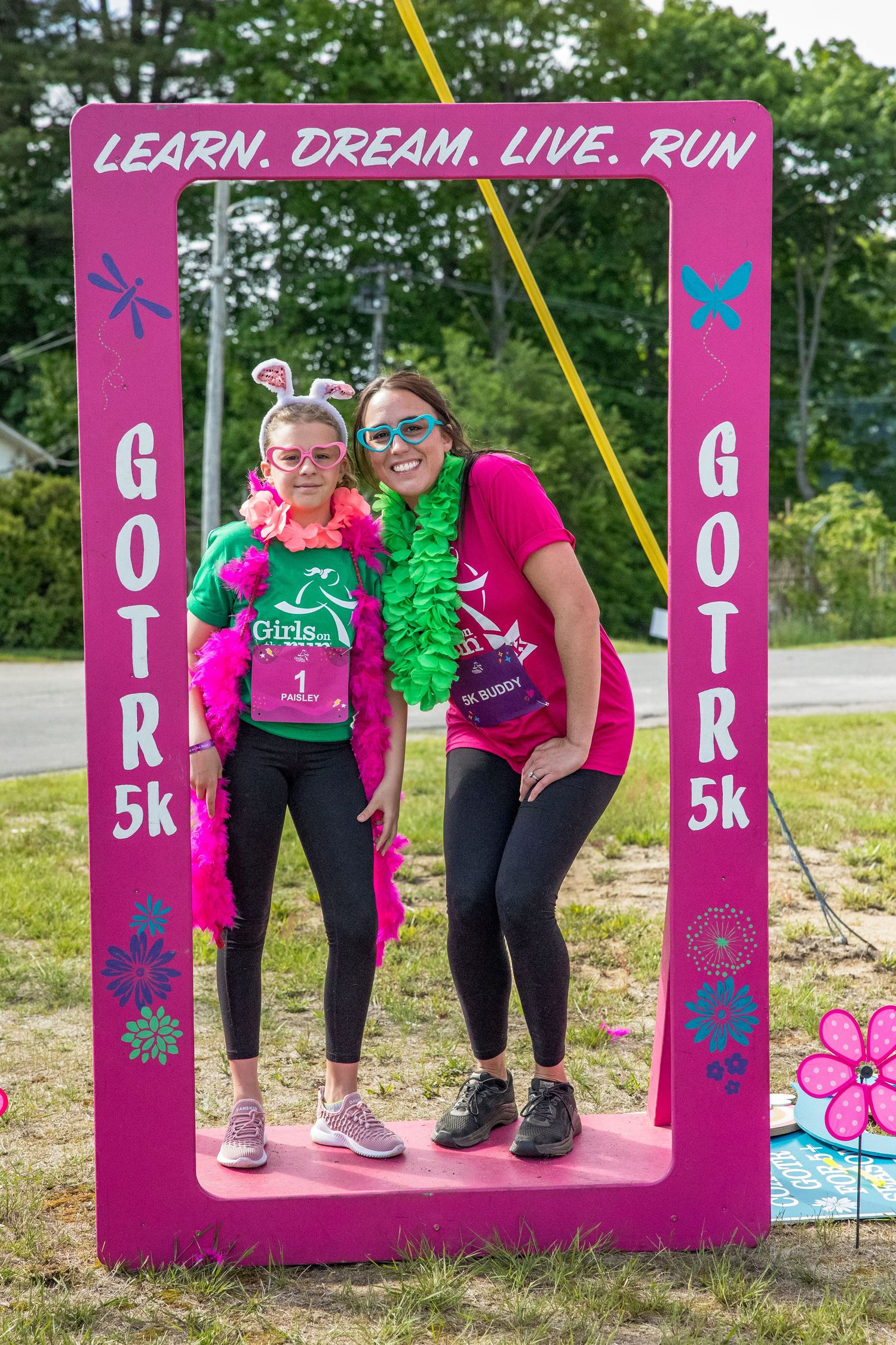 Two women pose in a pink GOTR 5K photo frame at a Girls on the Run event, wearing festive costumes outdoors.