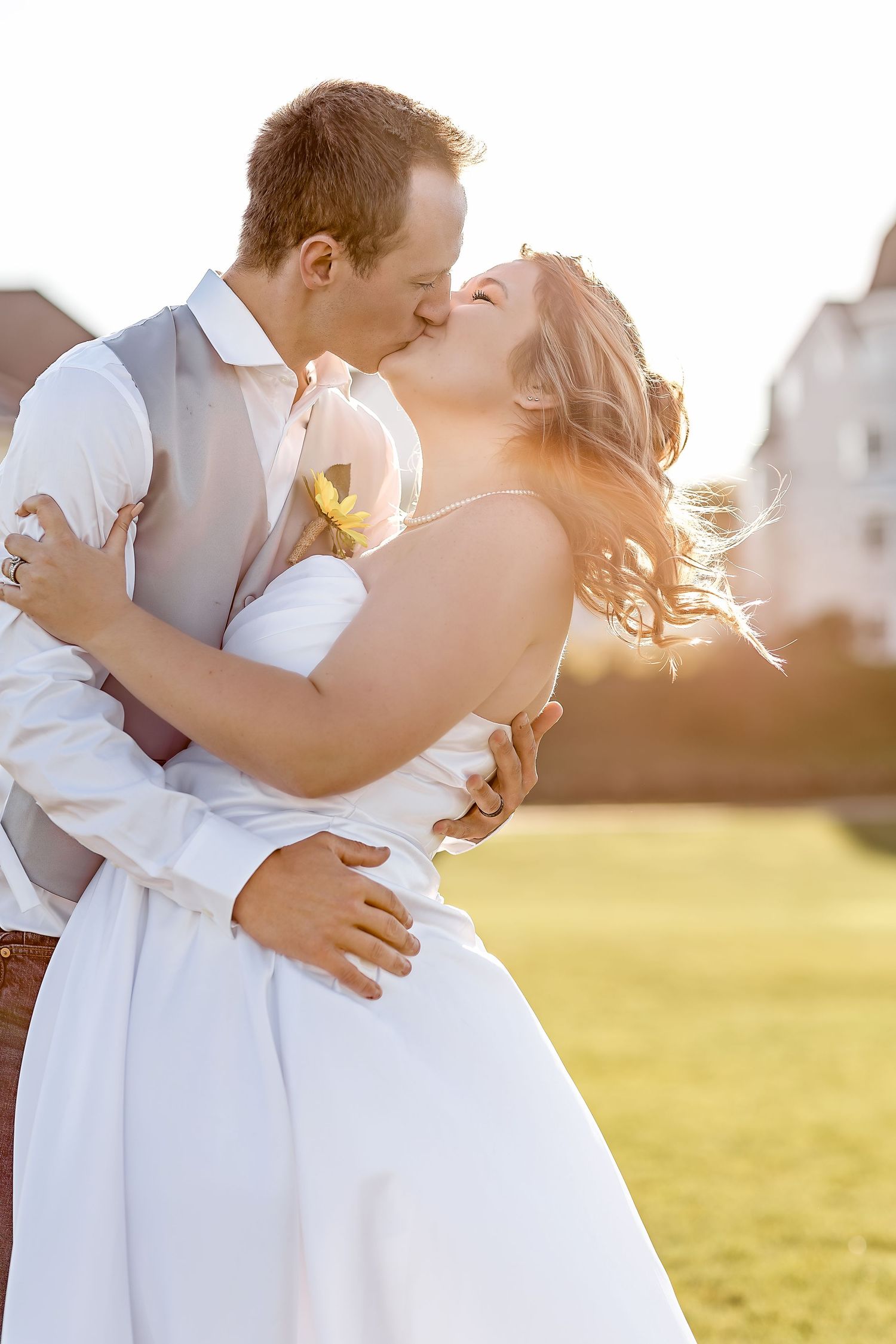 A bride and groom share a romantic kiss outdoors in golden sunlight, with the bride in a white gown and groom in a gray vest.
