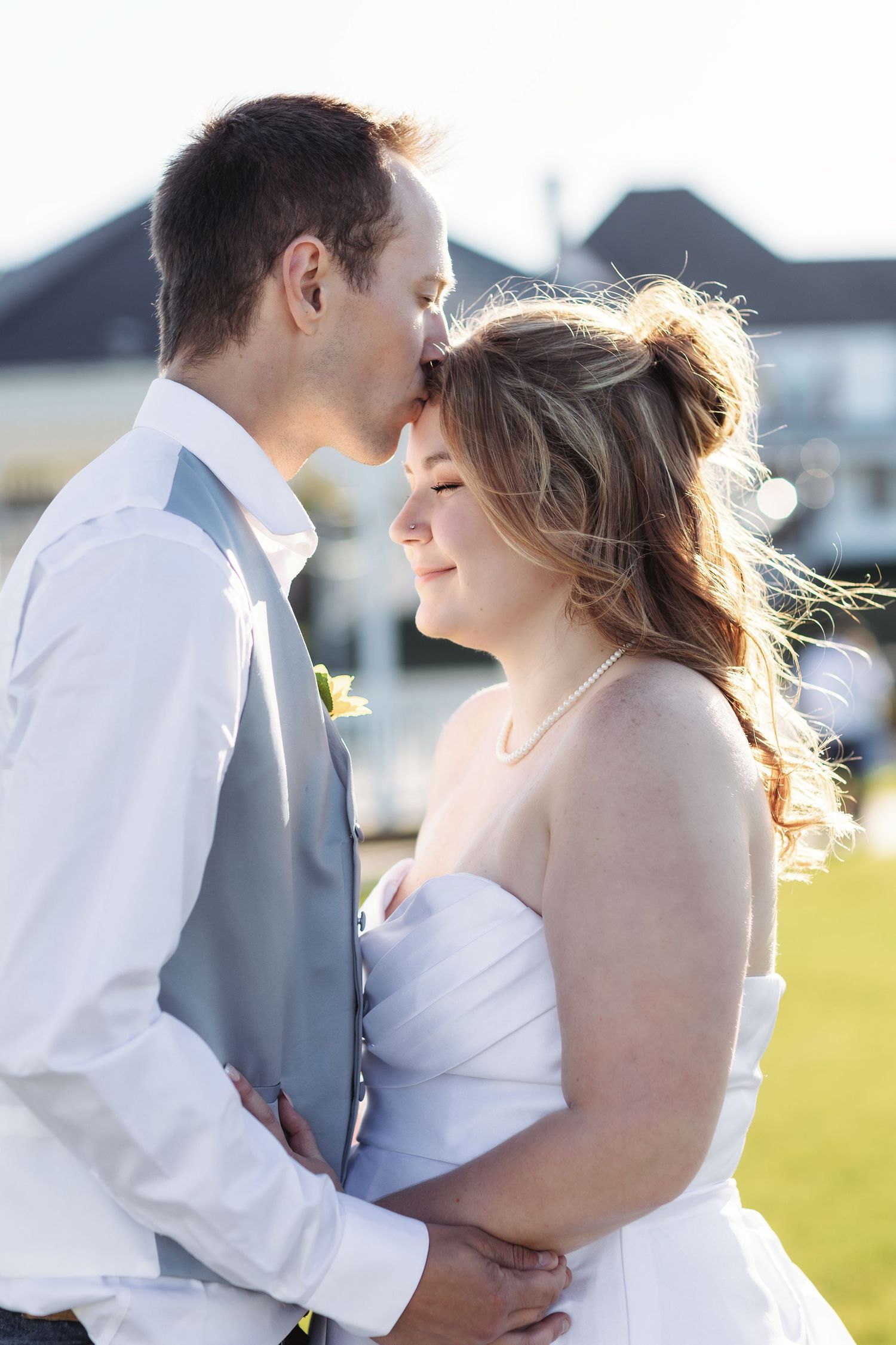 Bride and groom share a tender forehead kiss outdoors on their wedding day, with the bride smiling in a white strapless gown.