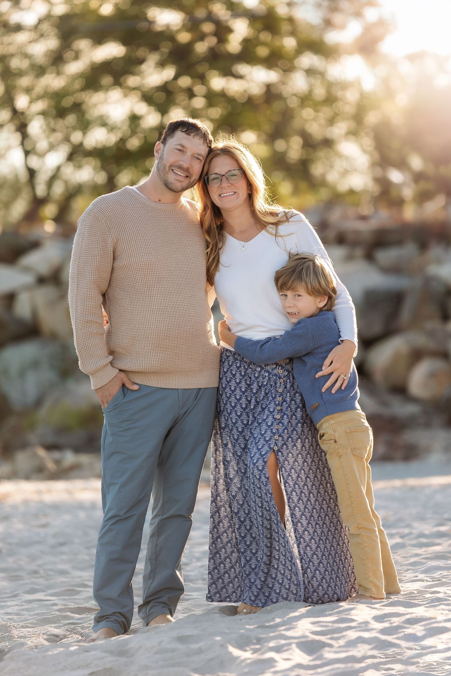 Family of three posing on sandy beach at golden hour, parents smiling with young boy between them.