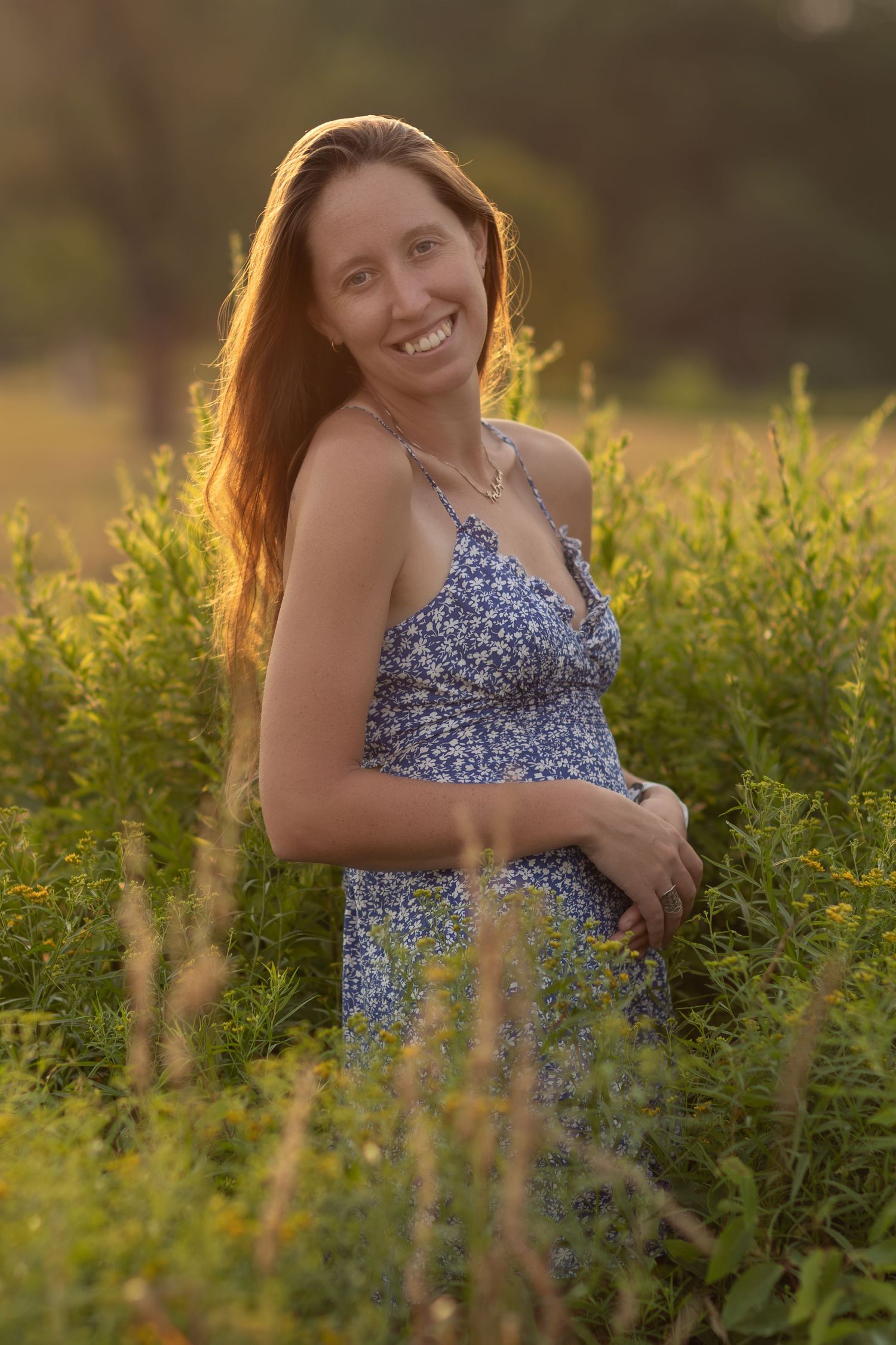 Smiling woman with long red hair in blue floral sundress standing among lush green plants in golden hour sunlight.