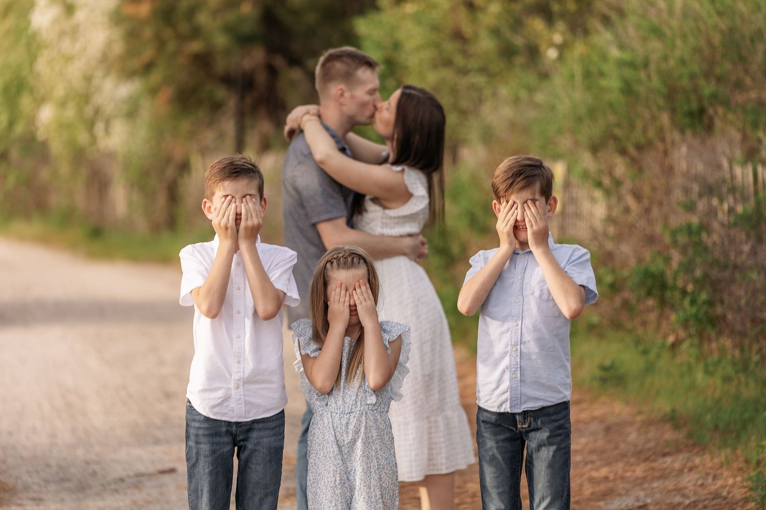 Family photo with parents kissing on a dirt path while three children cover their eyes in embarrassment outdoors.