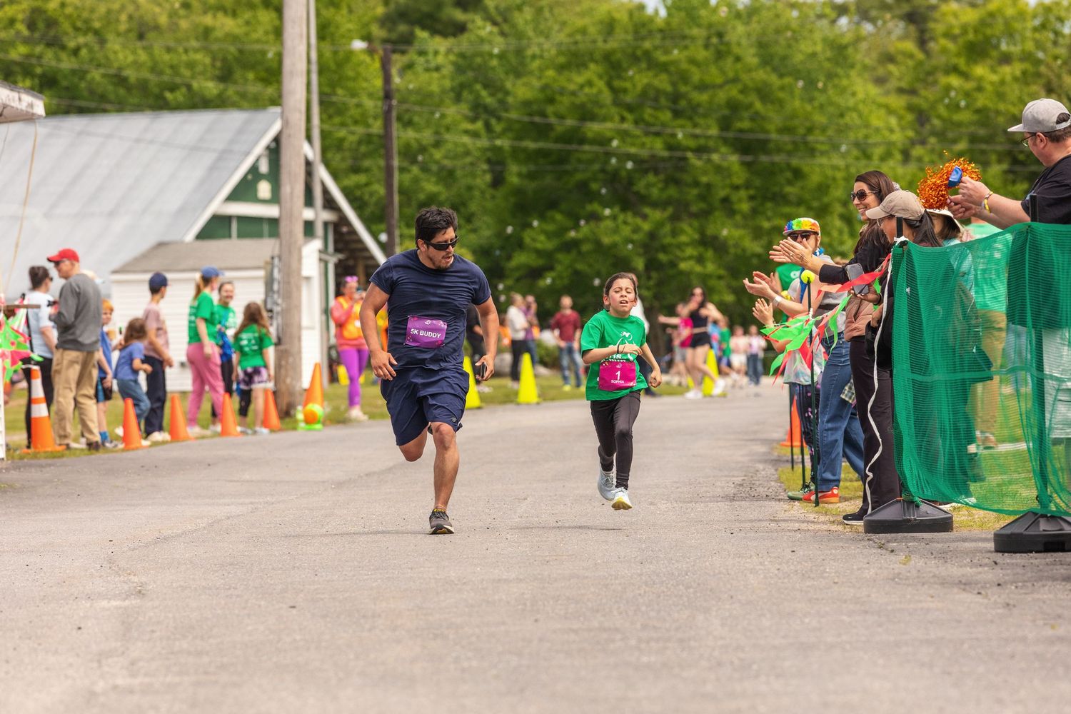 Runners cross the finish line at an outdoor charity race event with cheering spectators lining the course.