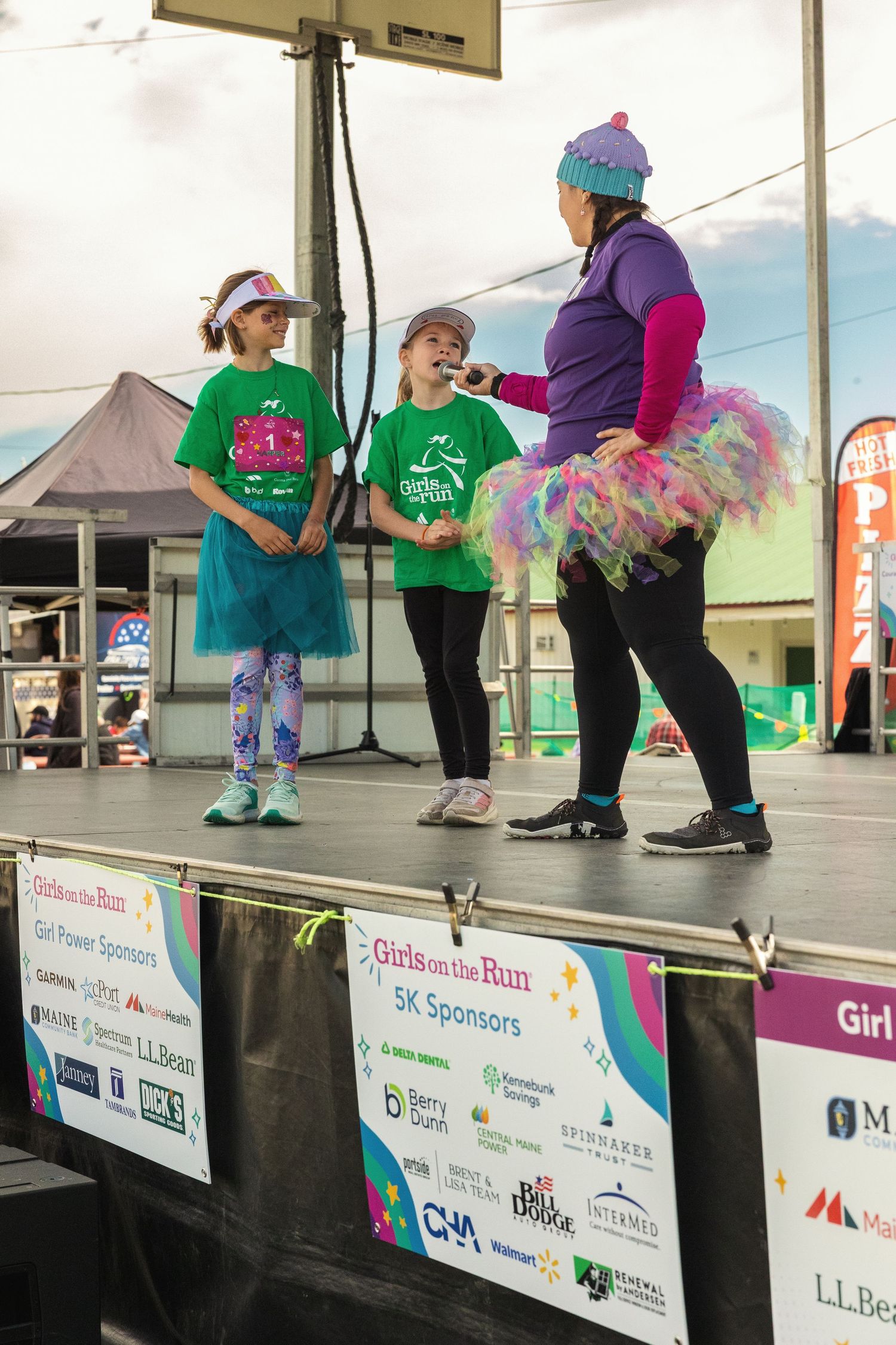Two girls in green Girls on the Run shirts stand on stage with a woman in a colorful tutu at a 5K event.