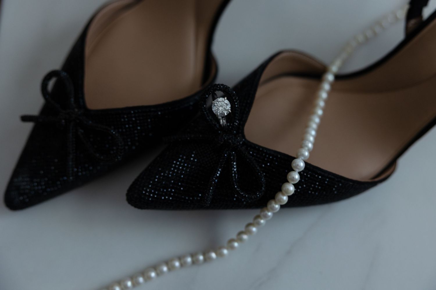Black glitter heels with pearl necklace accessories arranged in a repeating pattern against a neutral background. Wedding flat-lay.