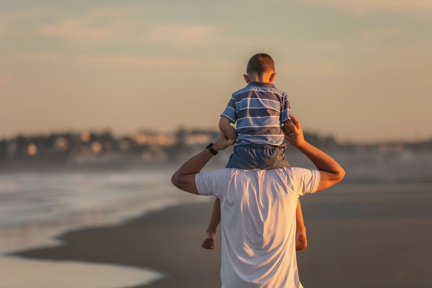 A father carries his young child on his shoulders while walking along a scenic beach at sunset.