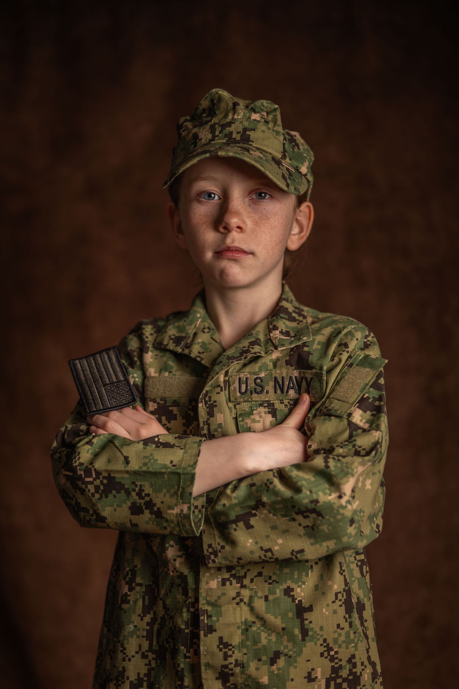 Young boy in U.S. Navy camouflage uniform and cap, arms crossed, posing seriously against dark background.