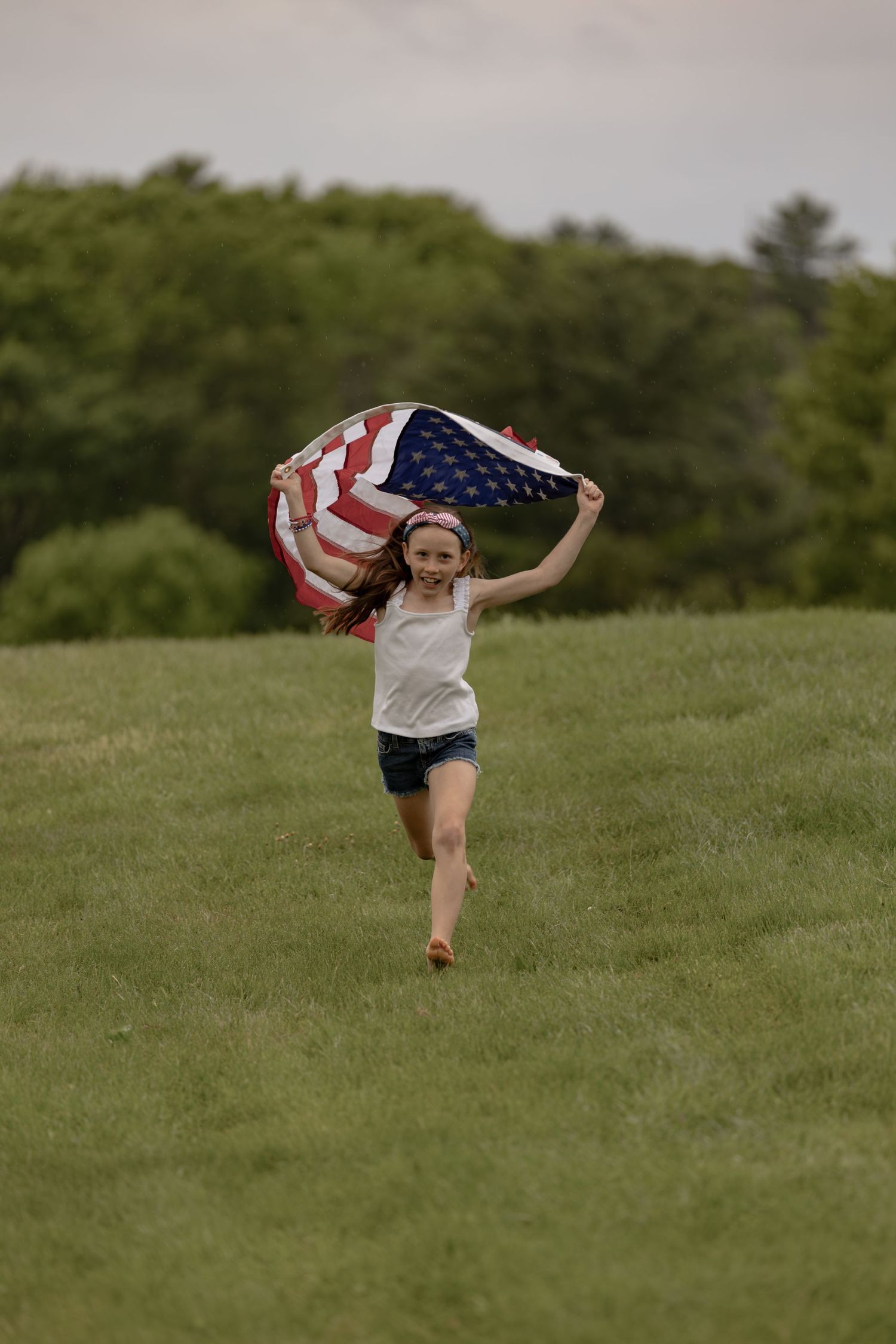 A joyful girl runs across a green field holding an American flag above her head on a cloudy day.