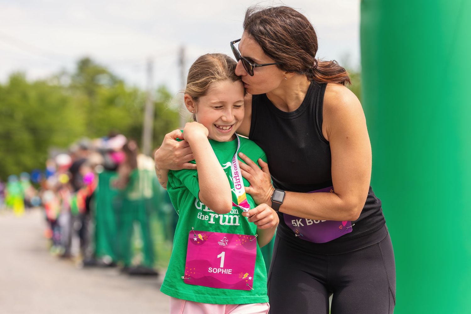 A woman hugs a smiling young girl wearing a race bib at an outdoor fun run event.
