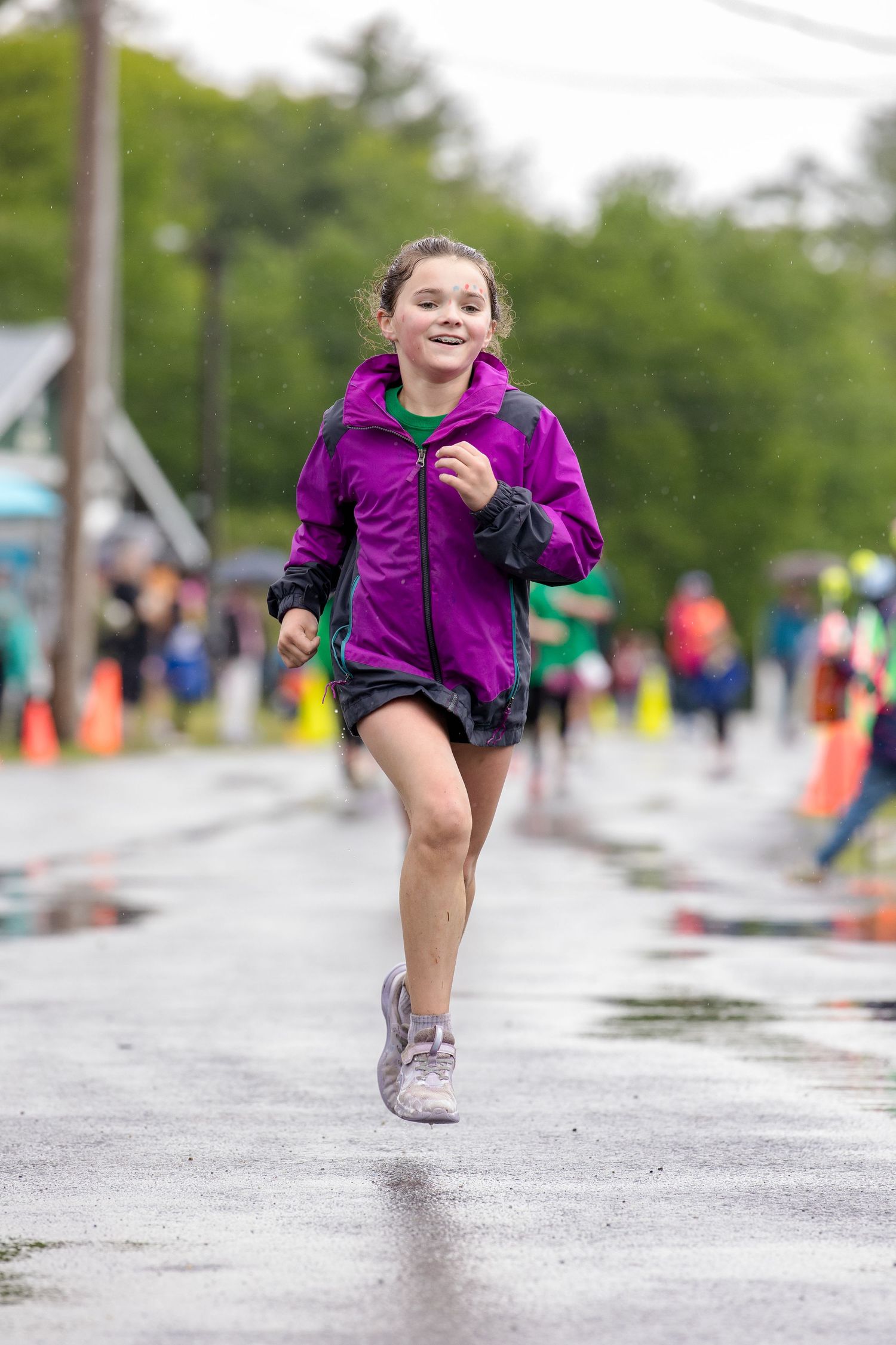 Young girl in purple jacket runs smiling on wet road during a race event with orange cones and spectators in background.
