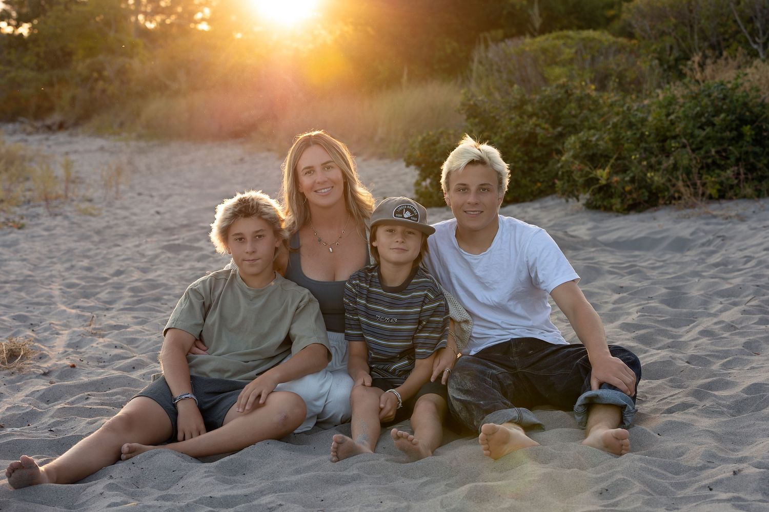 A family sits together on a sandy beach at sunset with golden sunlight glowing behind them.