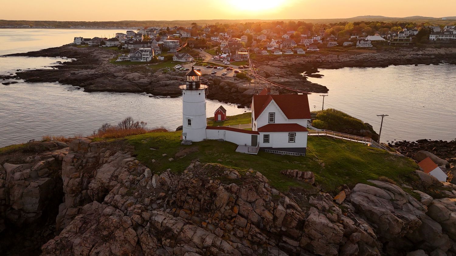 A lighthouse perched on rocky cliffs overlooks a coastal town at sunset, casting golden light across the harbor.
