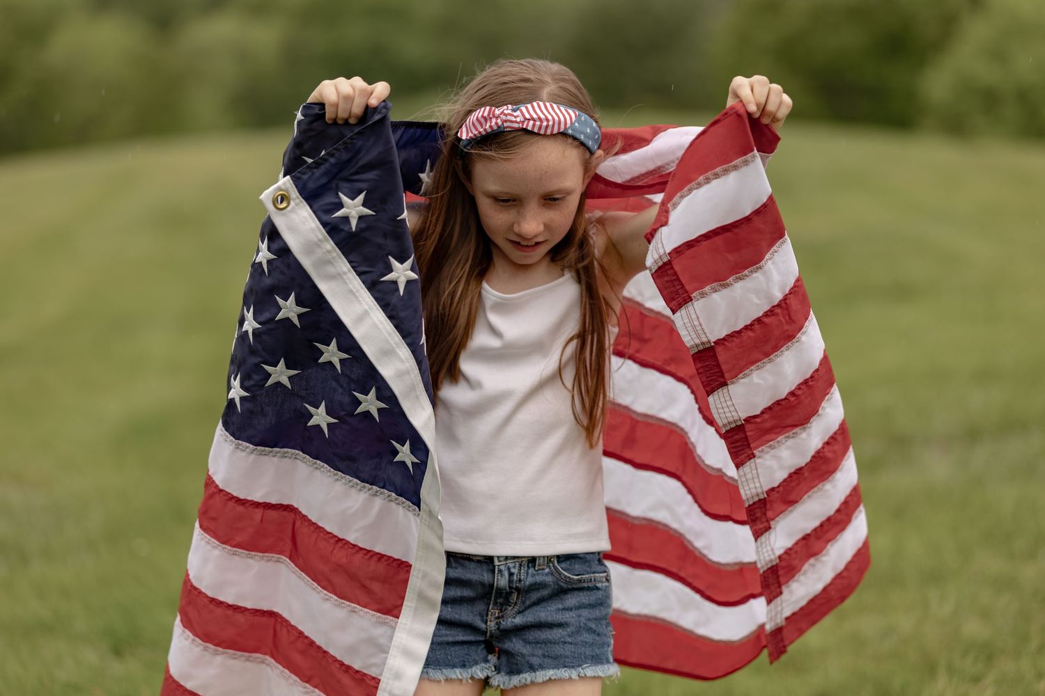 Young girl holding an American flag outdoors in a green field, wearing a white shirt and denim shorts.