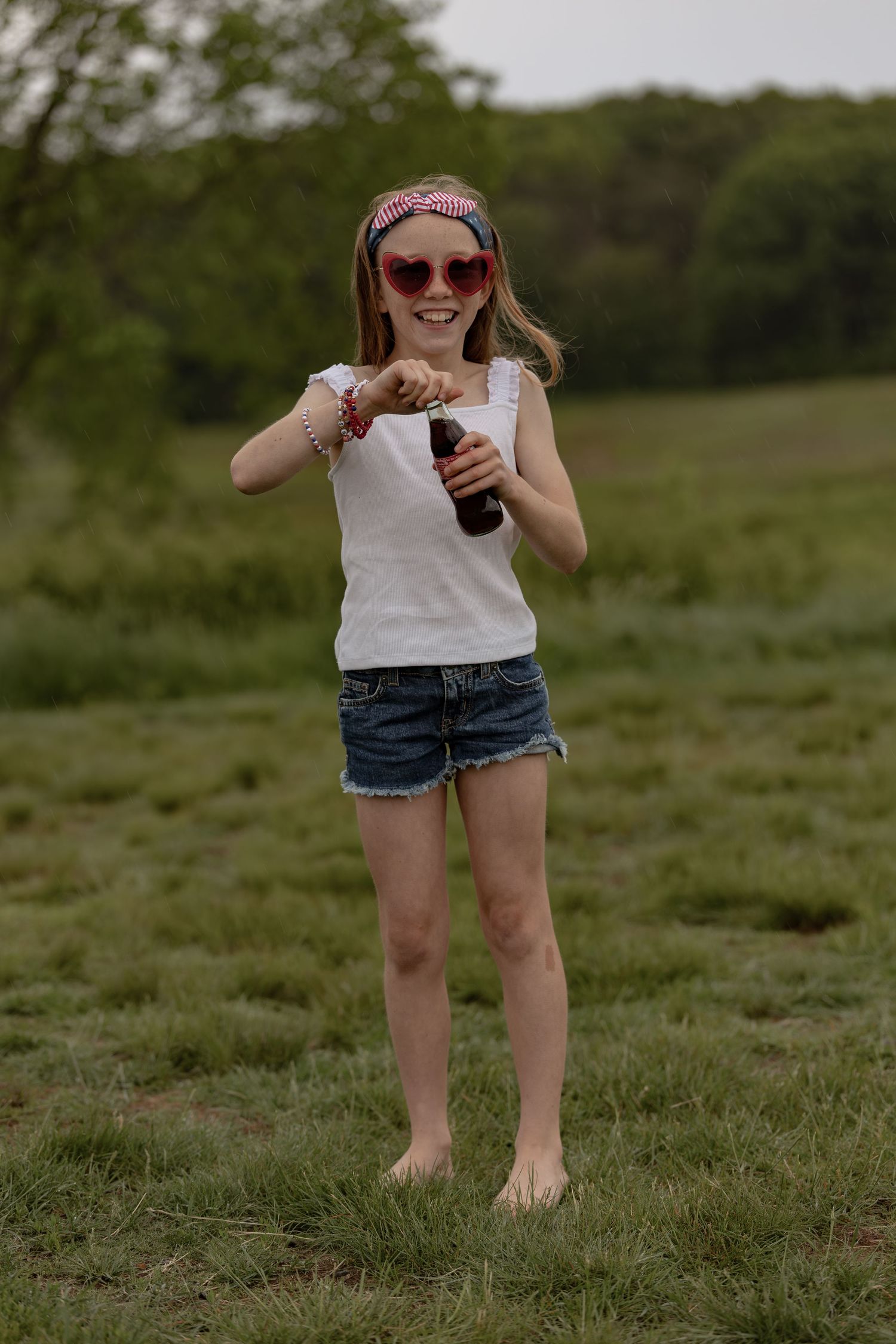 Young girl in heart sunglasses and denim shorts holds a soda bottle while standing barefoot in a green field.