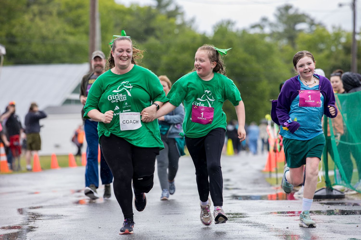Two women in green shirts joyfully running a 5K race on a wet road, smiling as they near the finish line.