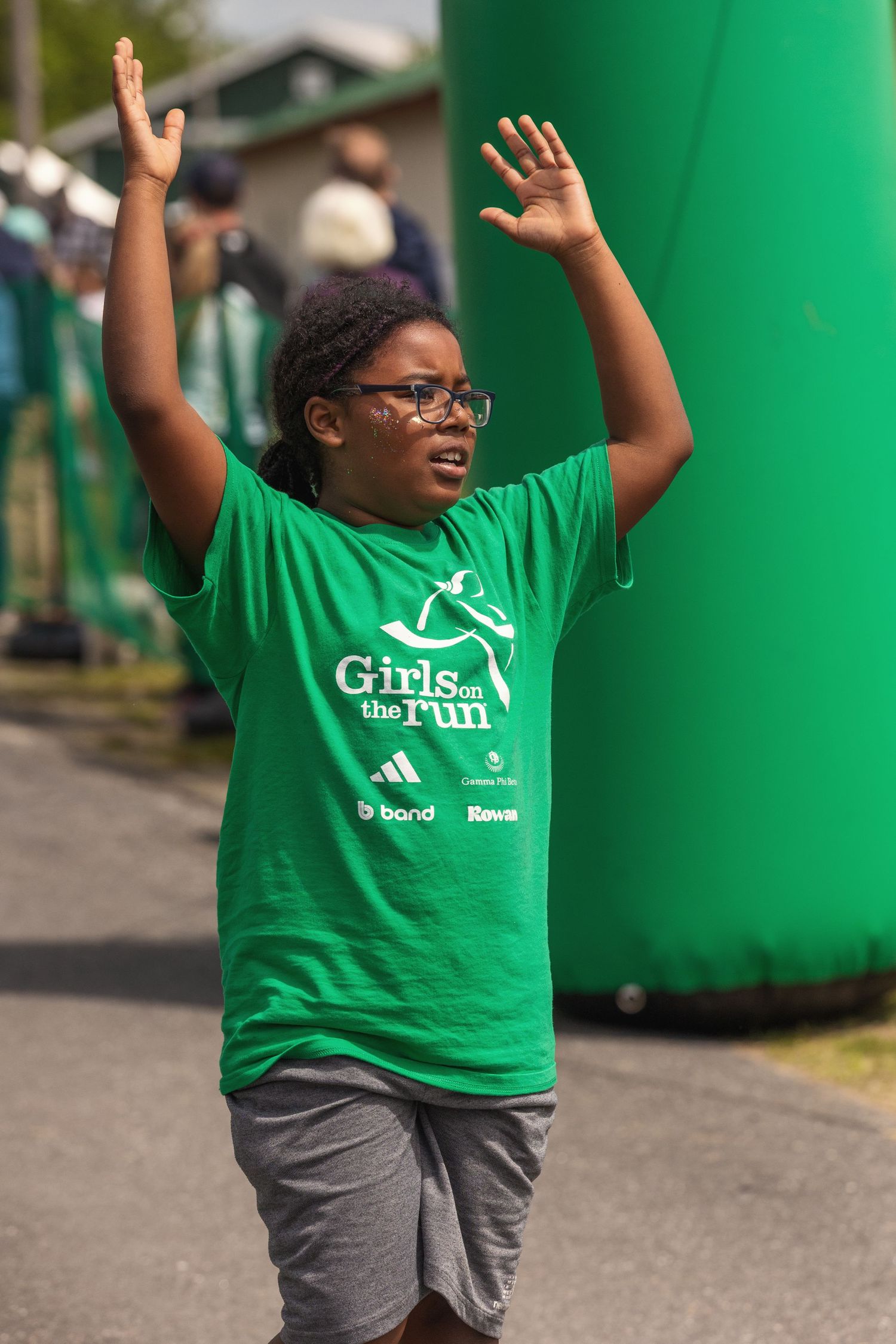 Young girl in green Girls on the Run t-shirt celebrates with arms raised at a charity fun run event.