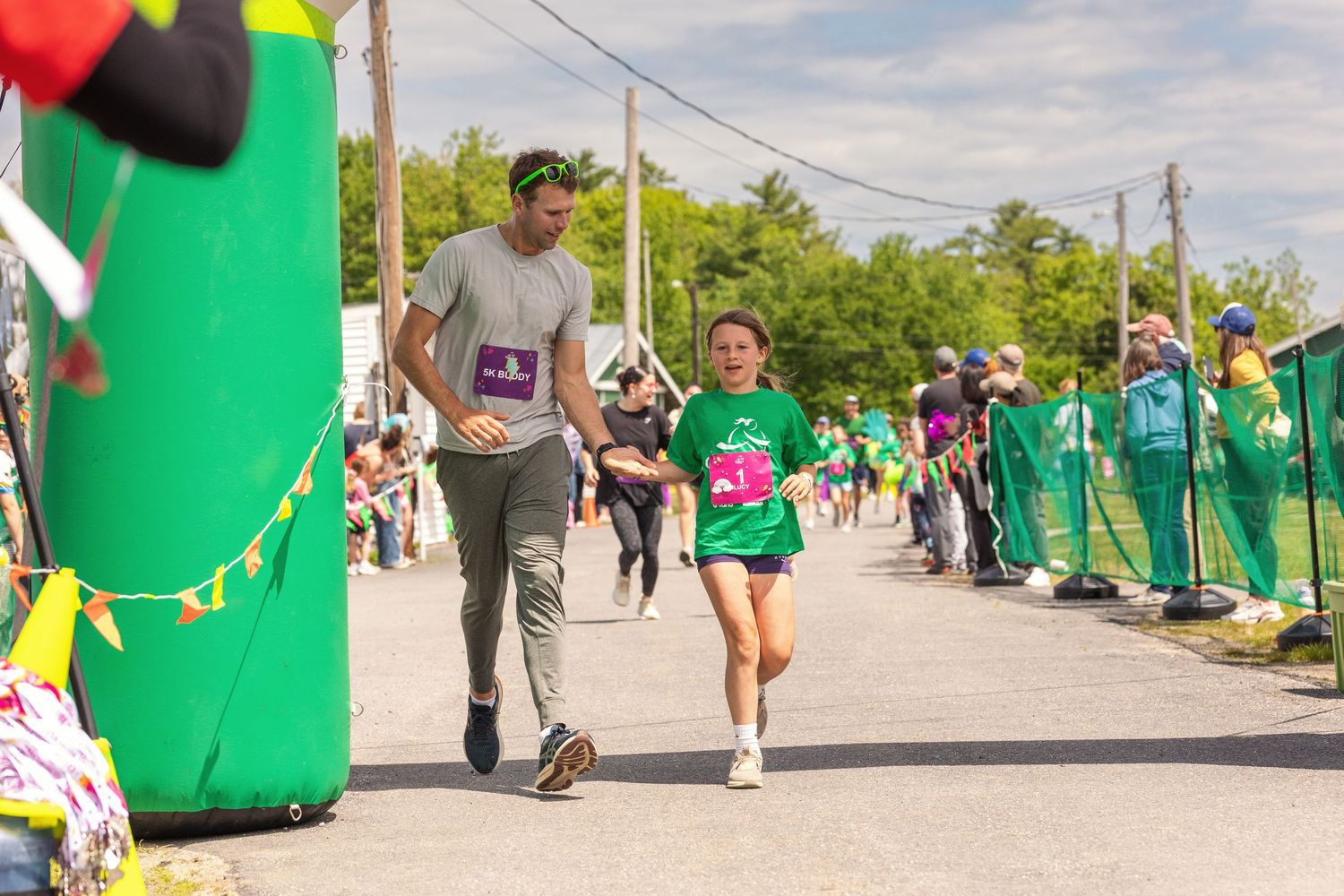 Adult and child running together in a fun outdoor charity race event with green decorations and spectators.