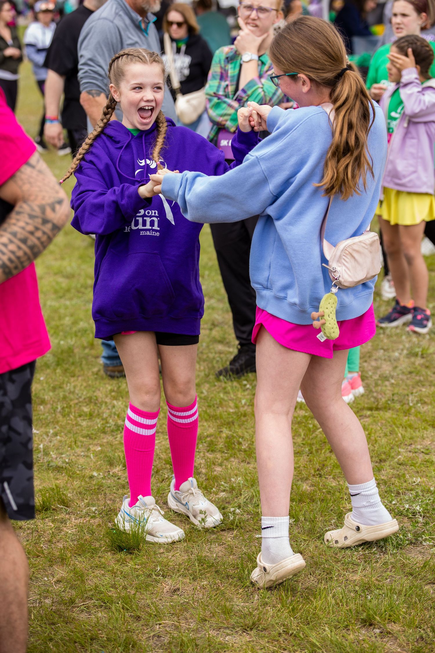 Two girls fist bumping at an outdoor event, wearing colorful hoodies and pink accessories on green grass.