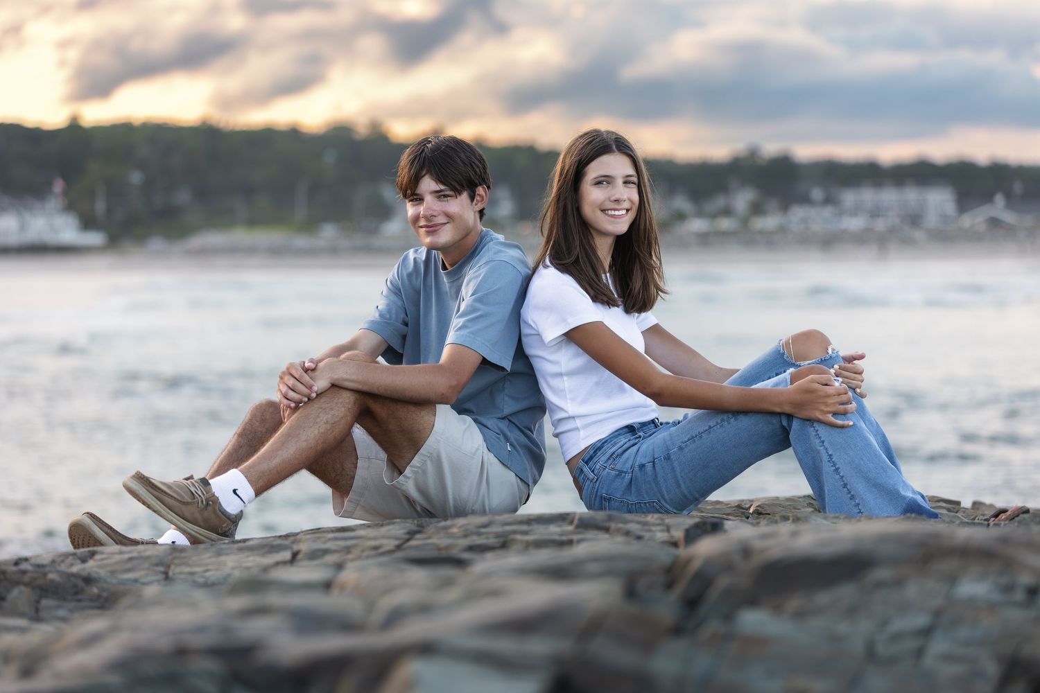 Two smiling friends sitting back-to-back on rocky shoreline at sunset, wearing casual jeans and white tops.