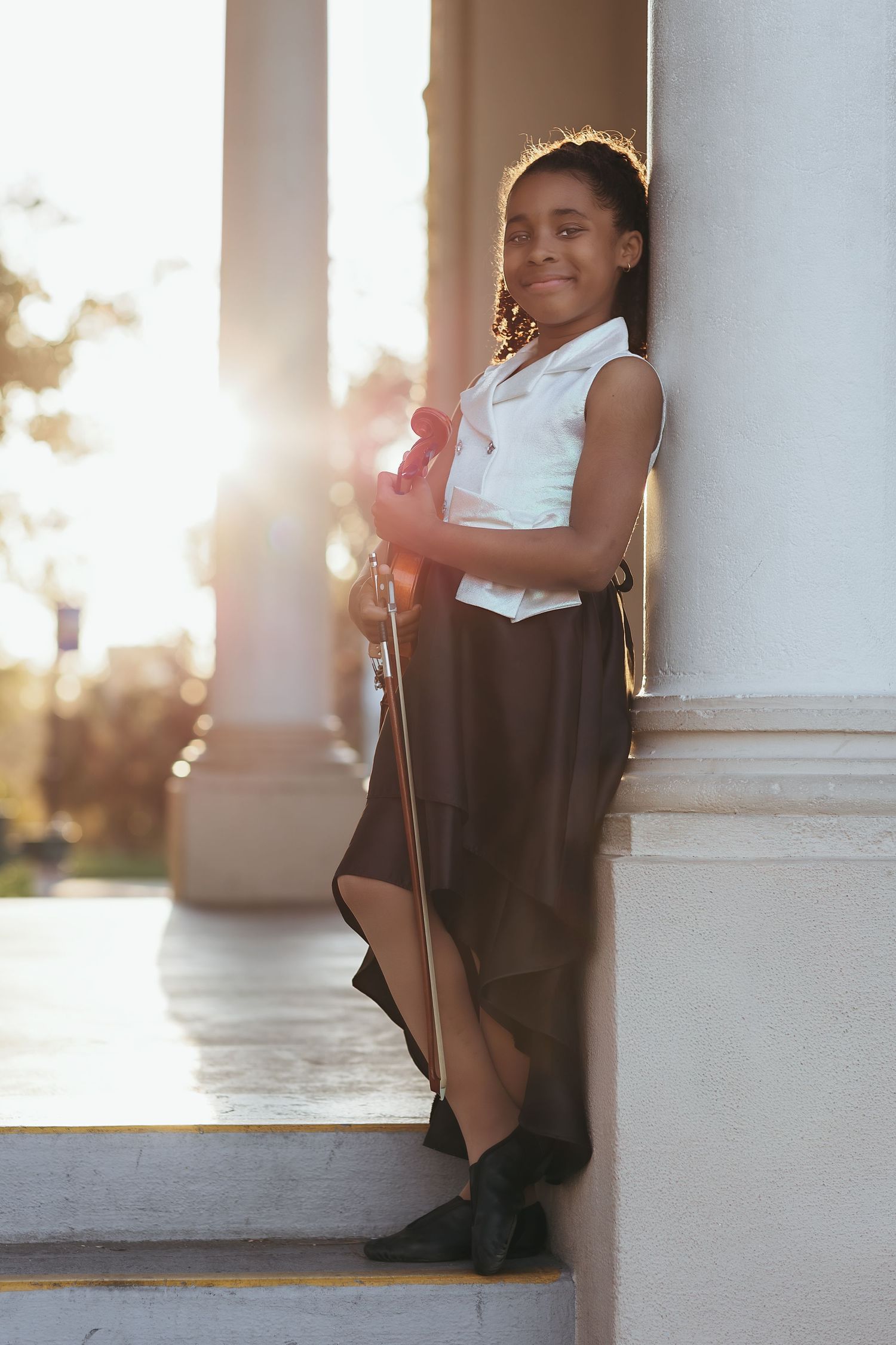 Young girl in white blouse and brown skirt leaning against a column holding a violin in golden sunlight.