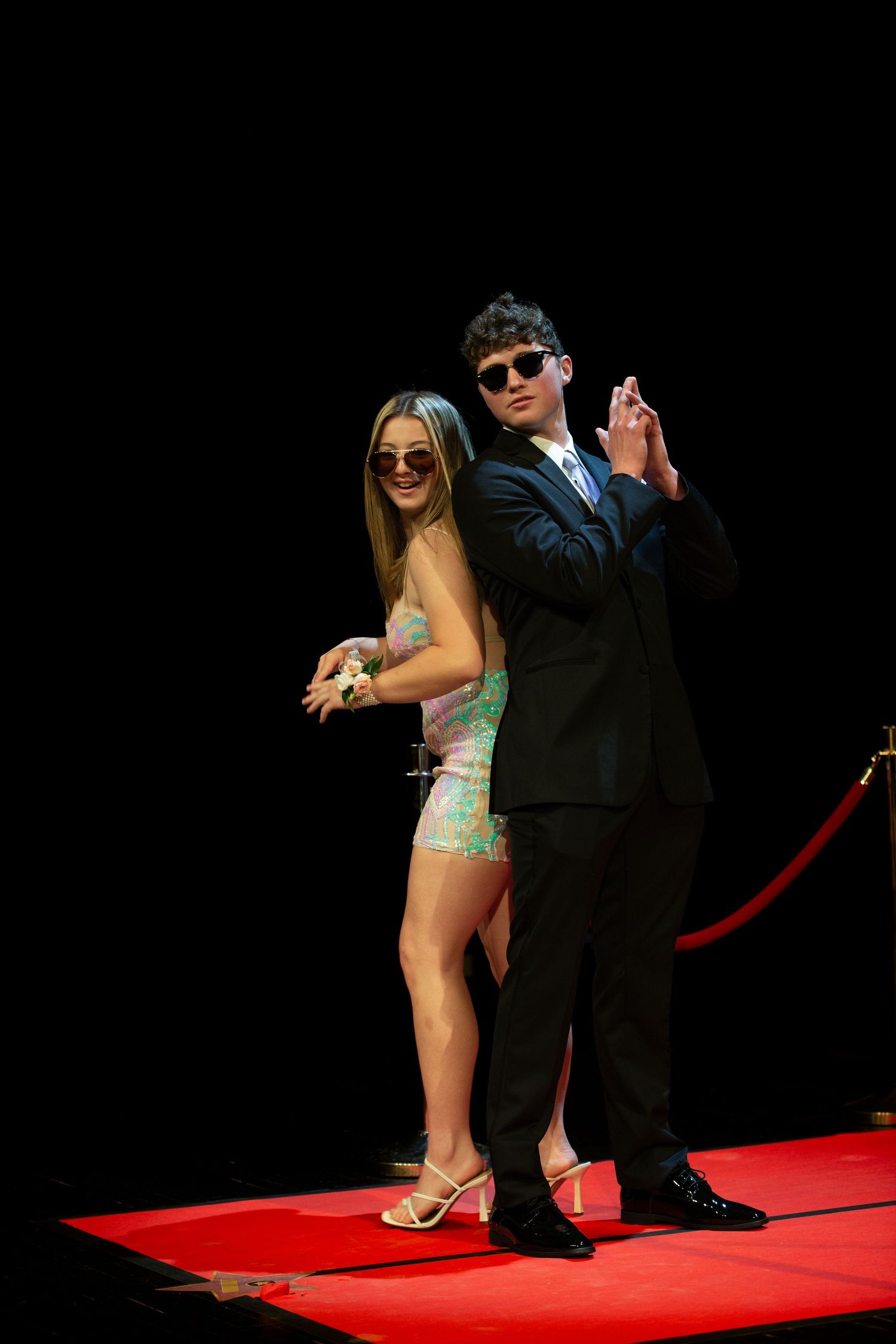 Young couple in formal attire posing on red carpet, woman in mini dress and man in black suit wearing sunglasses.