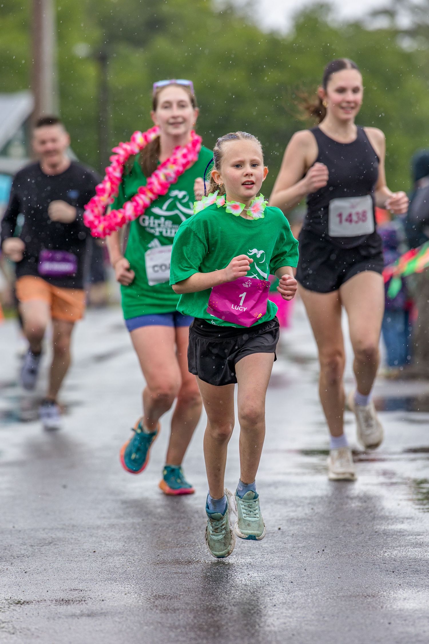 Young girl leads group of runners in rainy 5K race, wearing green shirt and pink bib number.