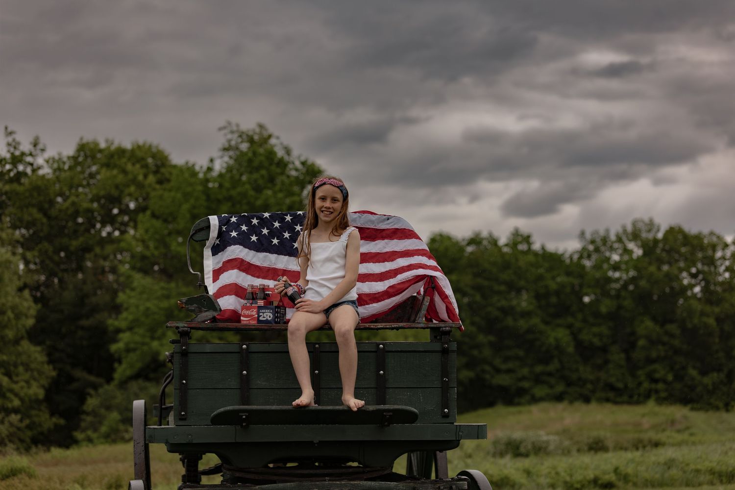 Girl sitting on vintage military vehicle with American flag on cloudy day in rural field.