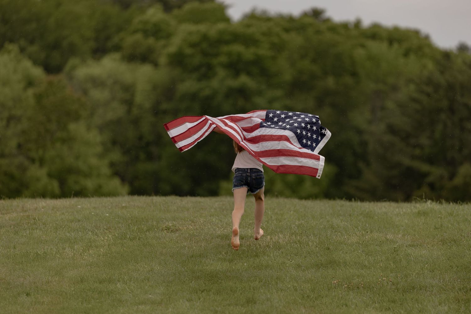Person running on green grass hill holding large American flag billowing in wind with trees in background.