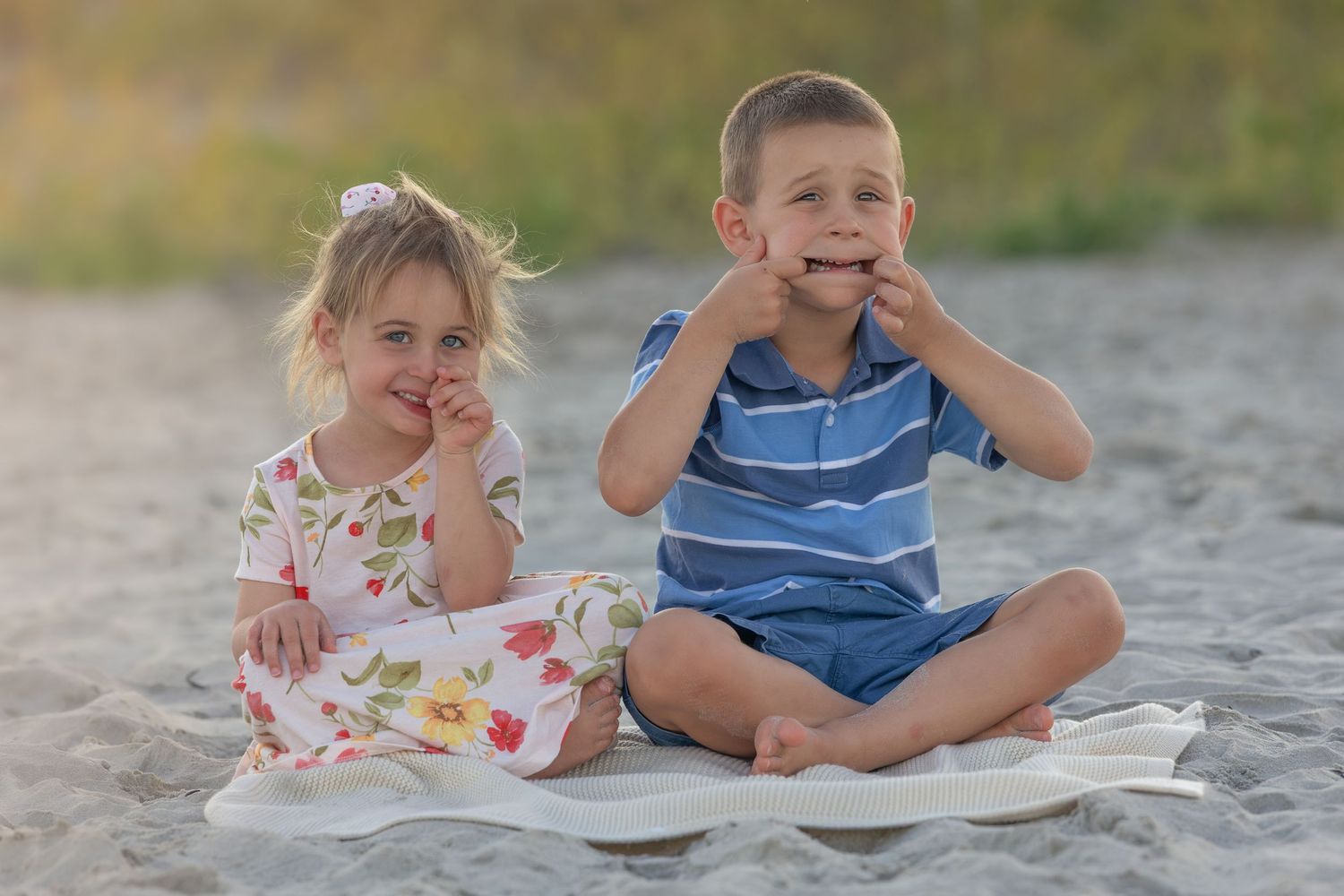 Two young children sitting on a blanket at the beach, smiling and eating snacks on a sunny day.