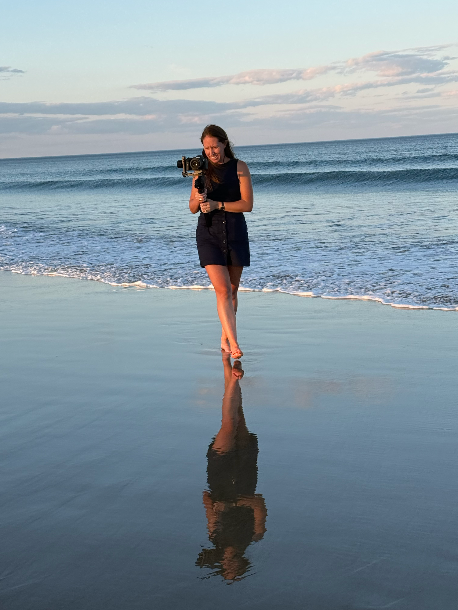Young woman holding a camera stands barefoot on a reflective wet beach at sunset with ocean waves behind her.