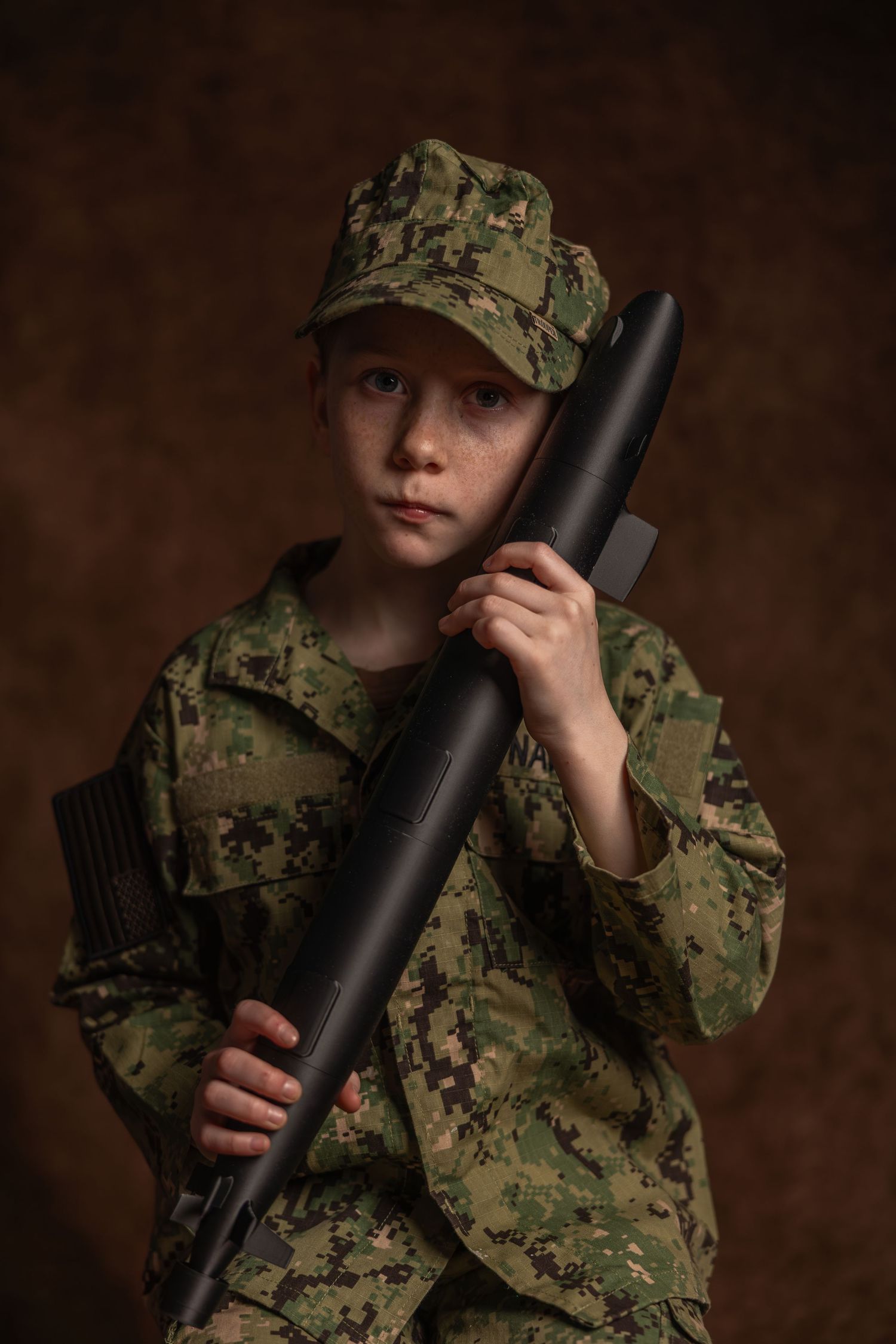 Young child in camouflage military uniform and cap holding a rifle prop in a studio portrait.