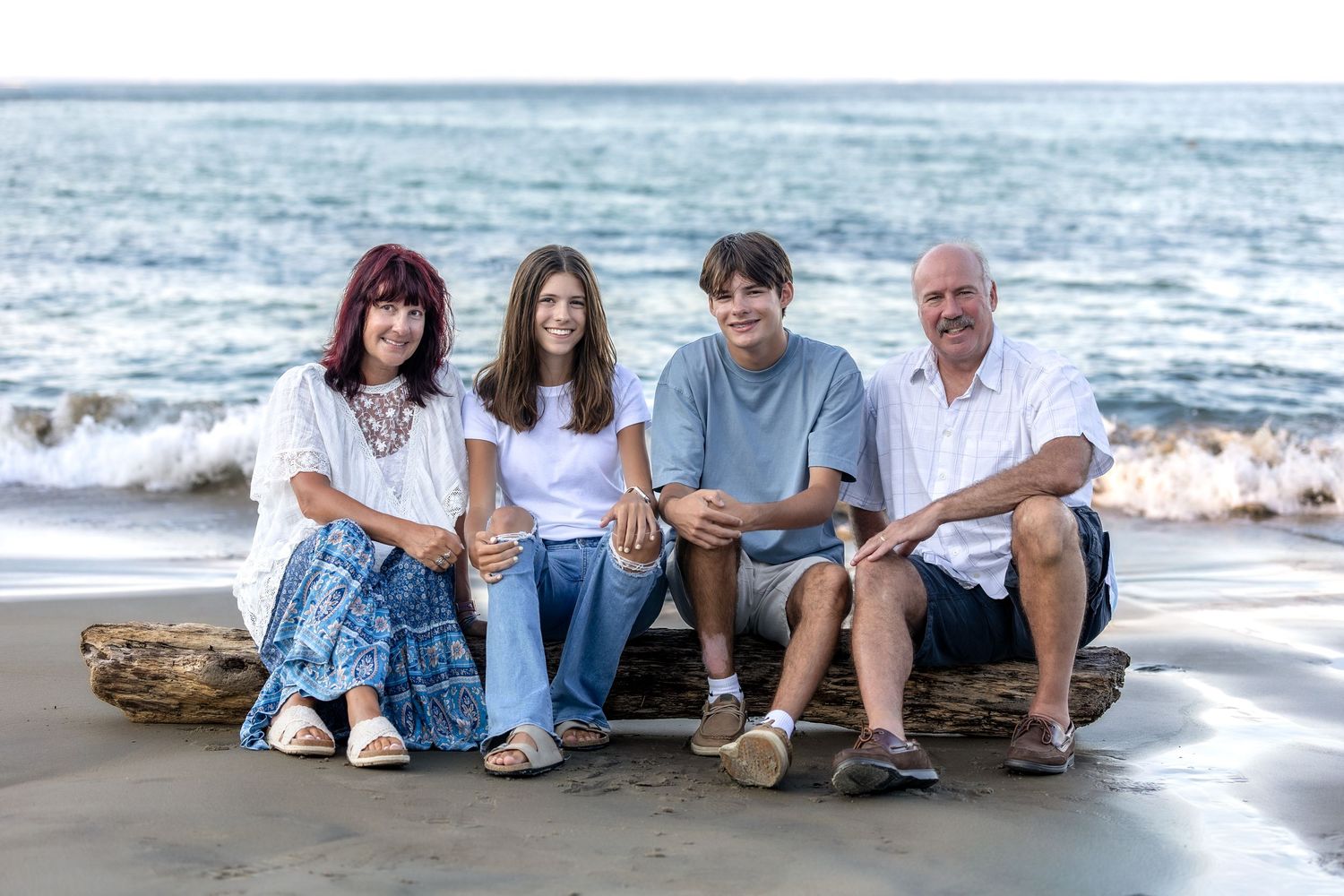 Family of four smiling together on a driftwood log at a sandy beach with ocean waves in the background.