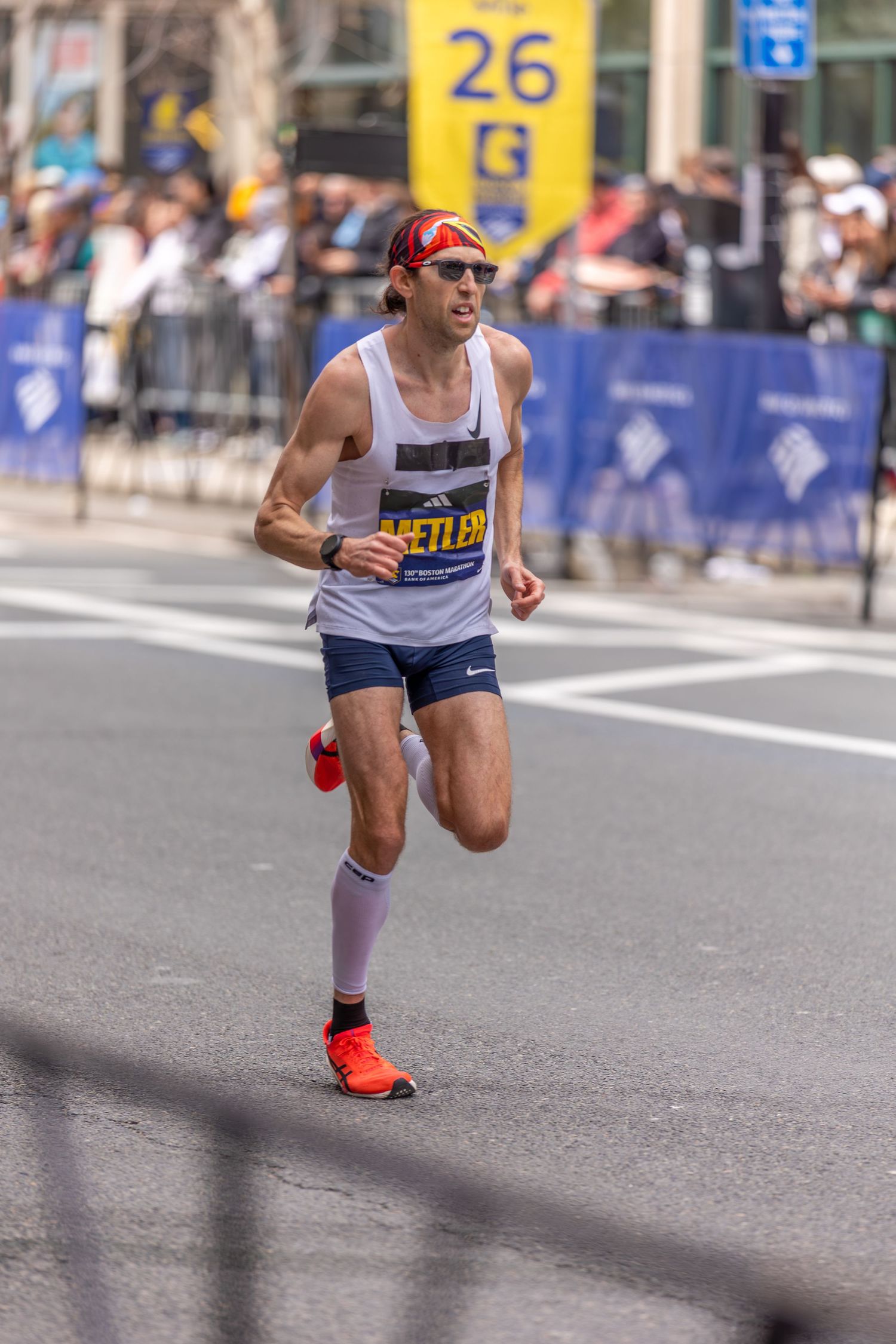 Male marathon runner named Shetler competing in a road race near mile marker 26, wearing sunglasses and orange shoes.
