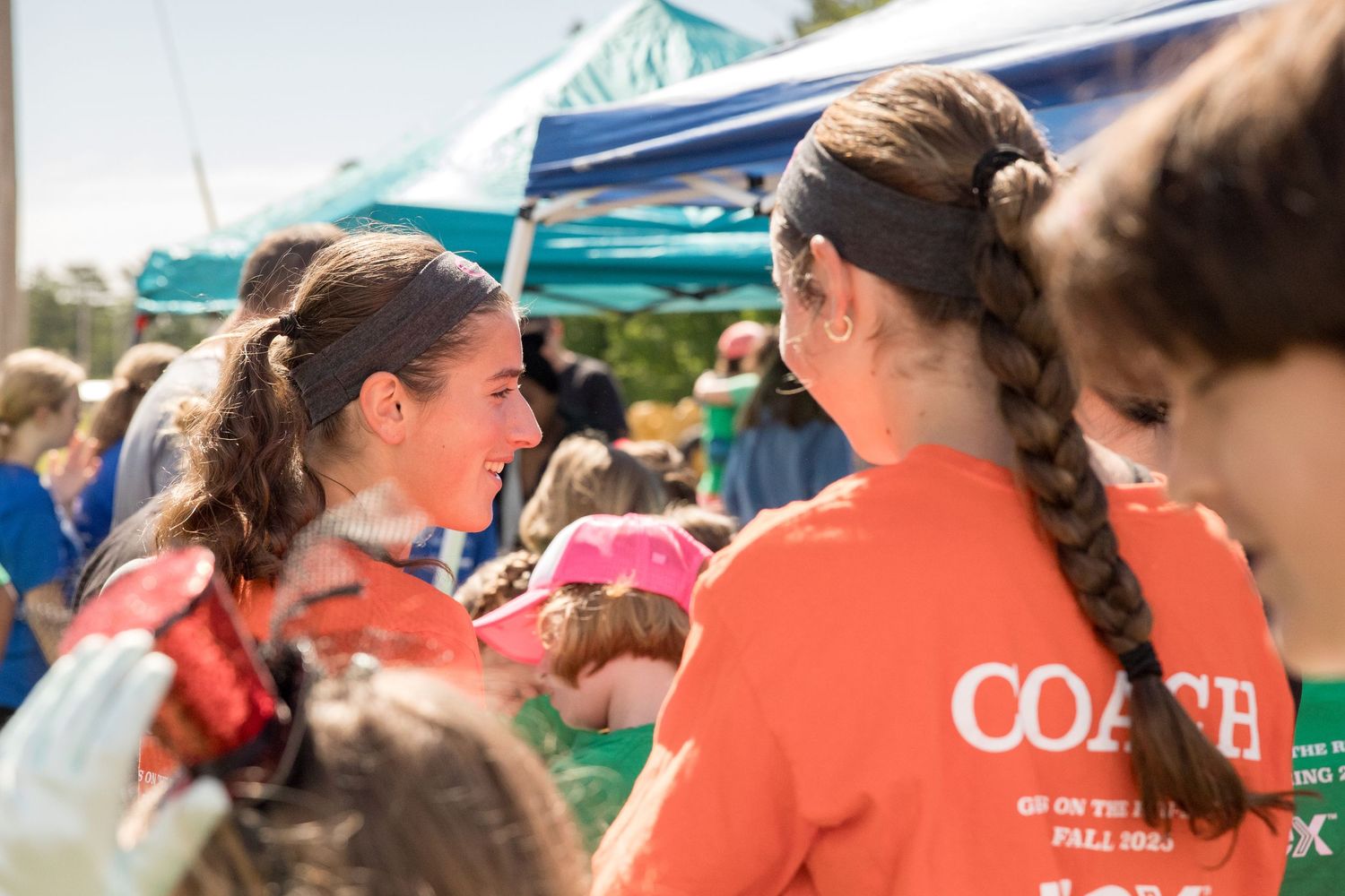 Two girls in orange Coach shirts chat at an outdoor event under a teal tent on a sunny day.