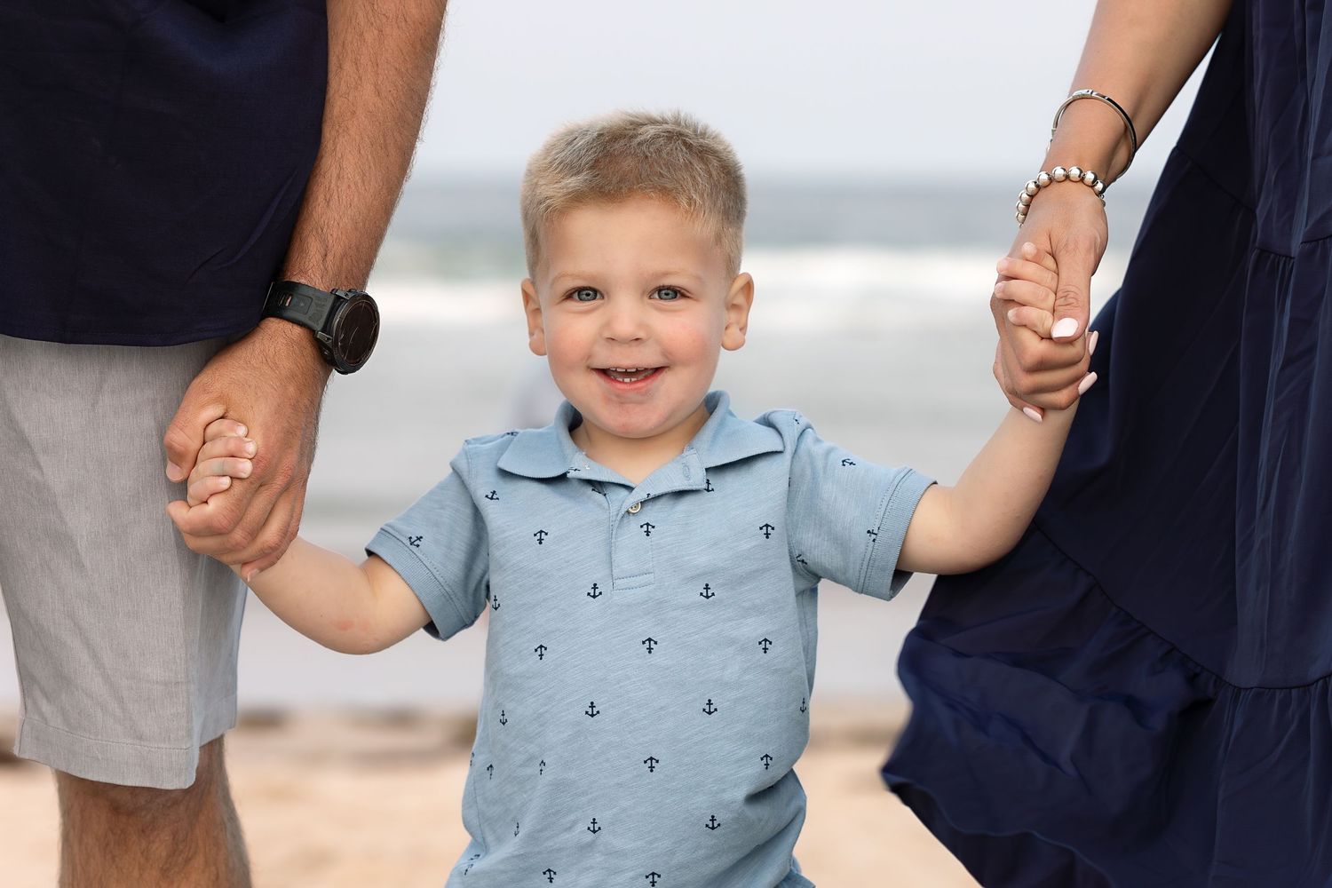 Happy toddler boy in blue polo shirt holds parents hands on sandy beach with ocean waves in background.