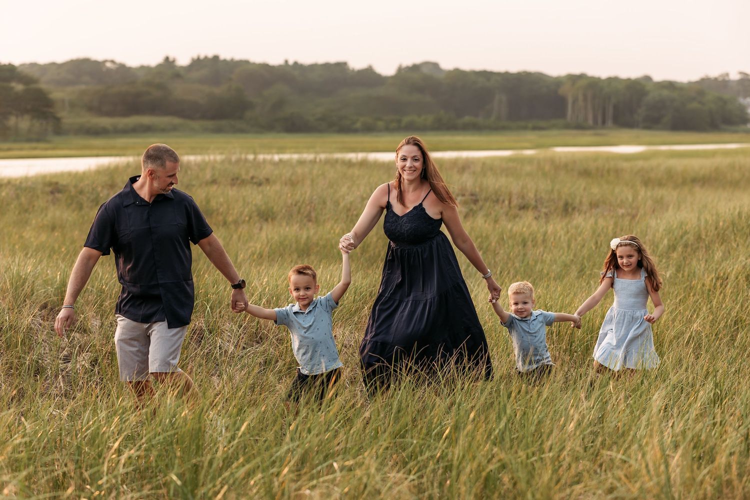 Family of five walking through tall grass near a marsh, parents holding hands with three young children outdoors.