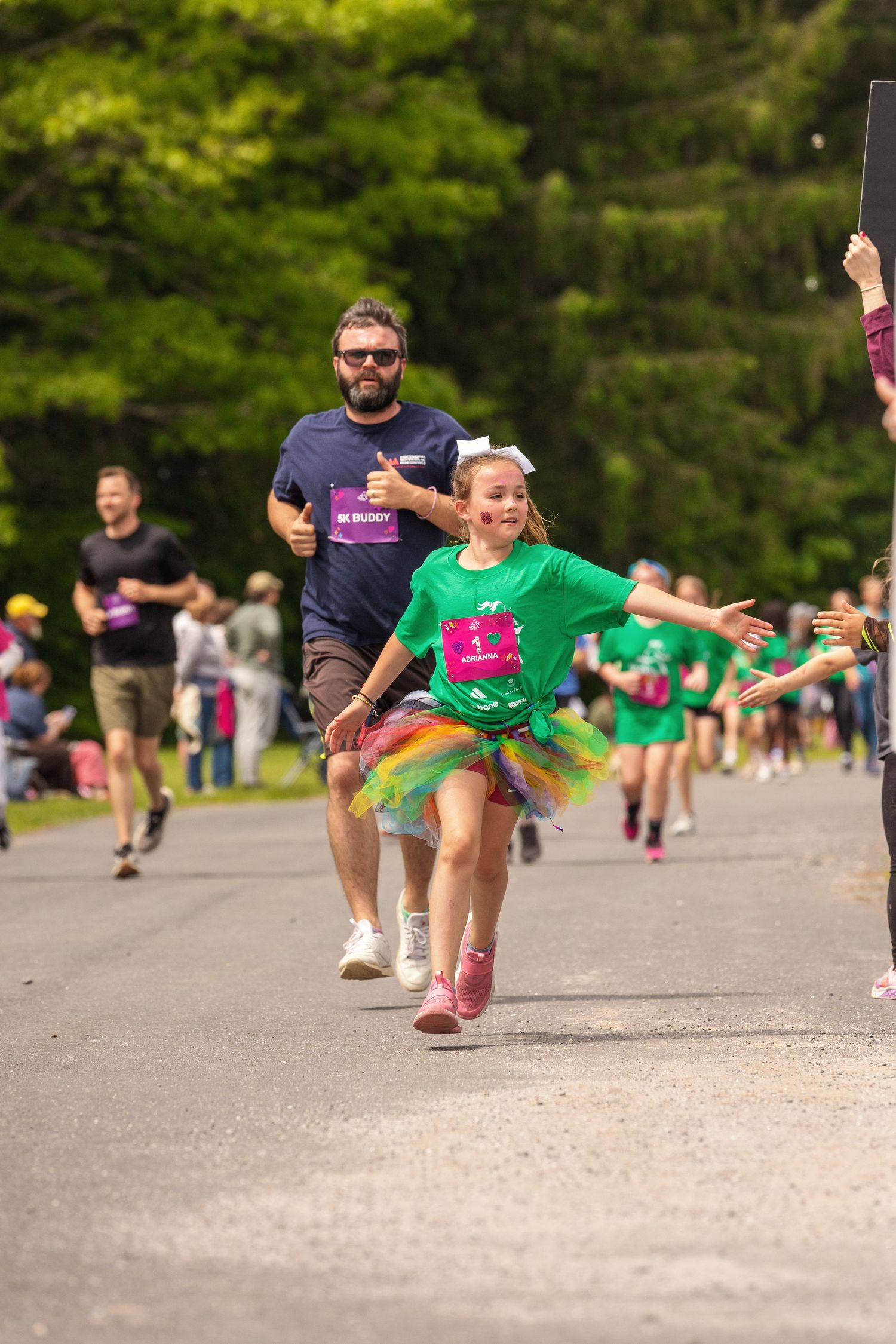 Girl in rainbow tutu and green shirt races toward finish line with bearded man behind her at charity fun run.