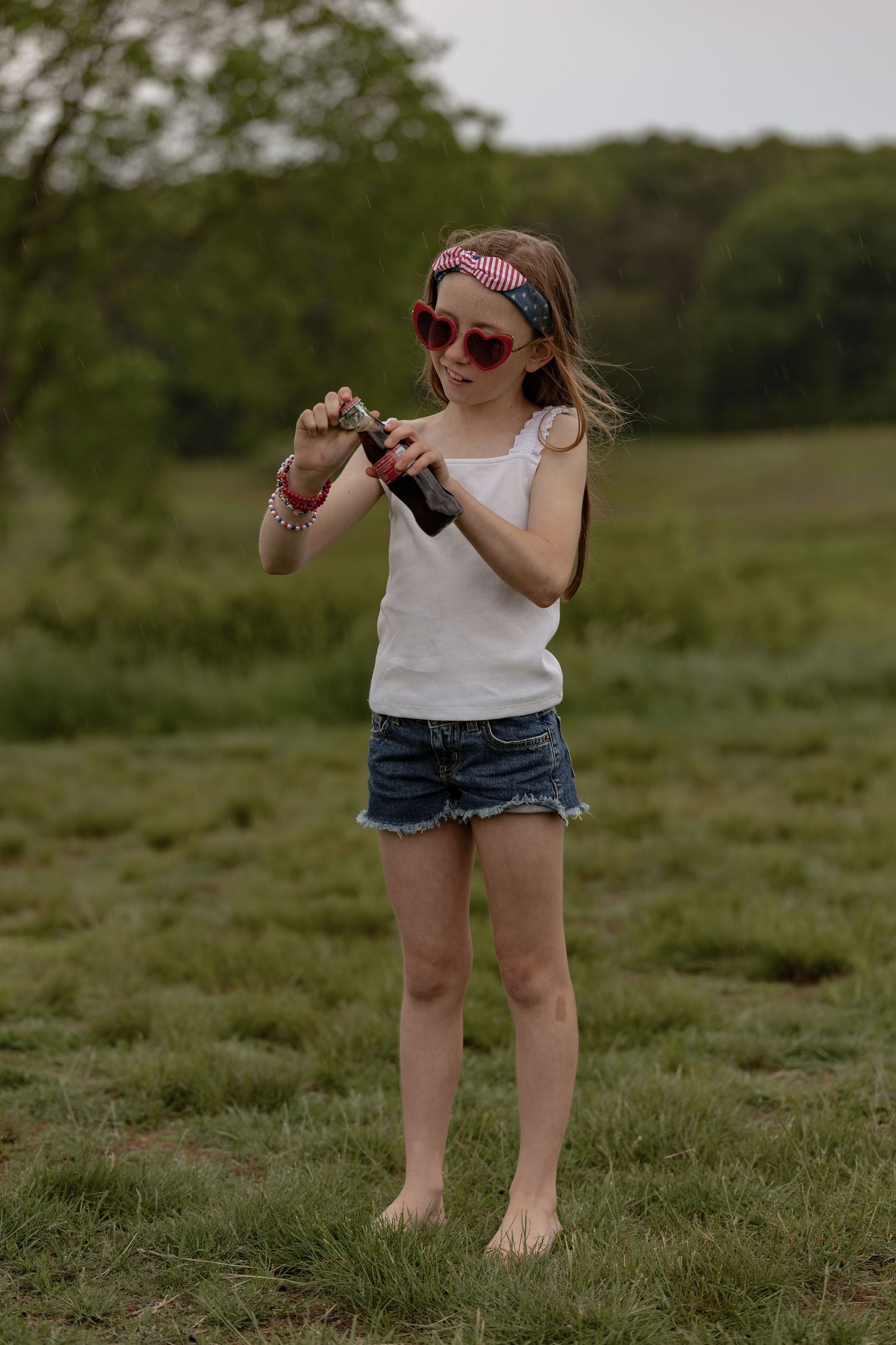 Young girl in white tank top, denim shorts, and heart sunglasses stands barefoot in green field holding a bottle.