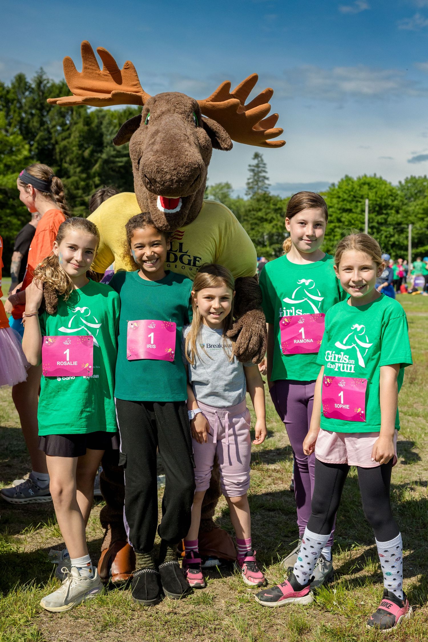 Girls on the Run participants pose with a moose mascot at an outdoor event, wearing green shirts and pink race bibs.