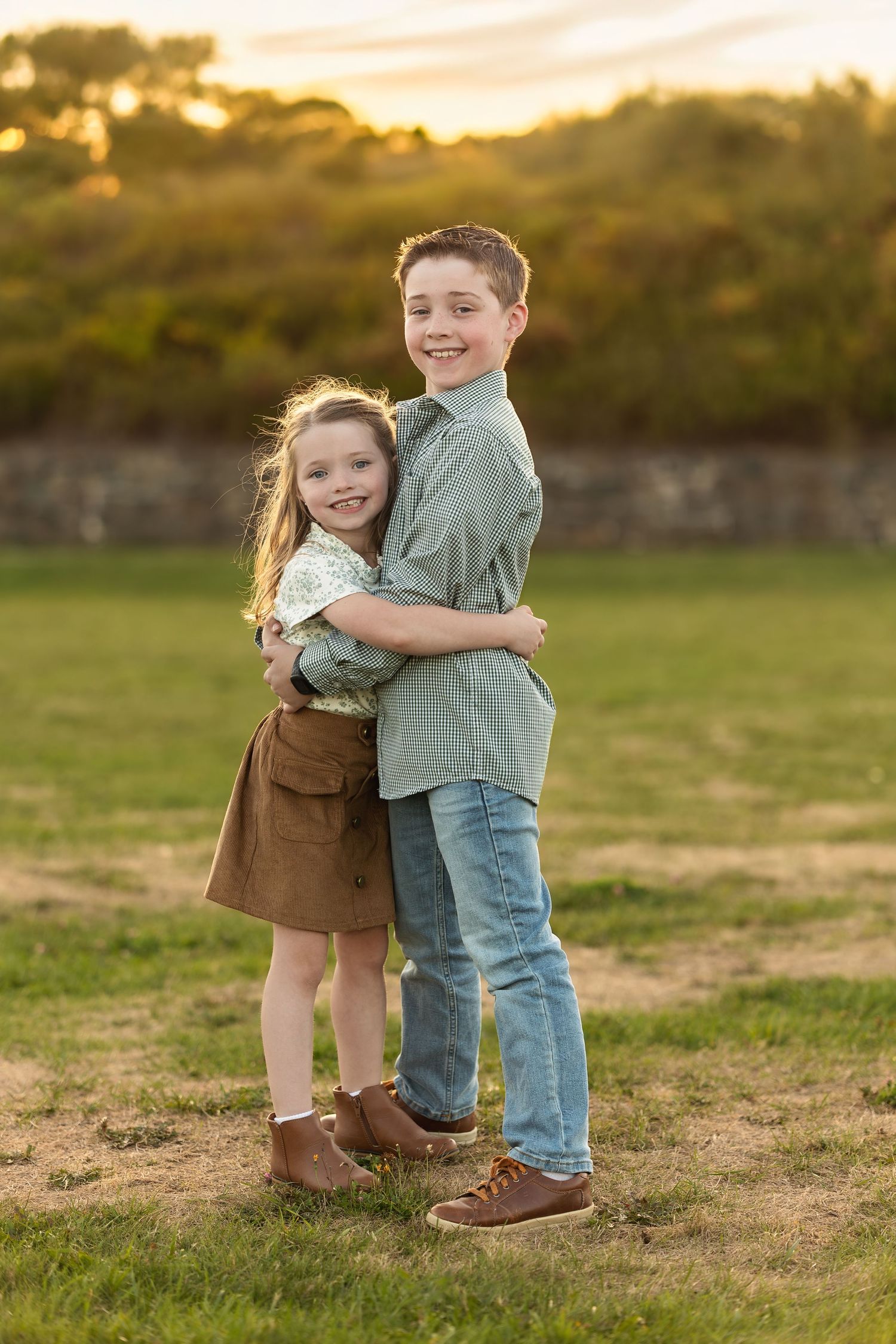 Two young siblings share a sweet hug while posing in a grassy field at sunset.