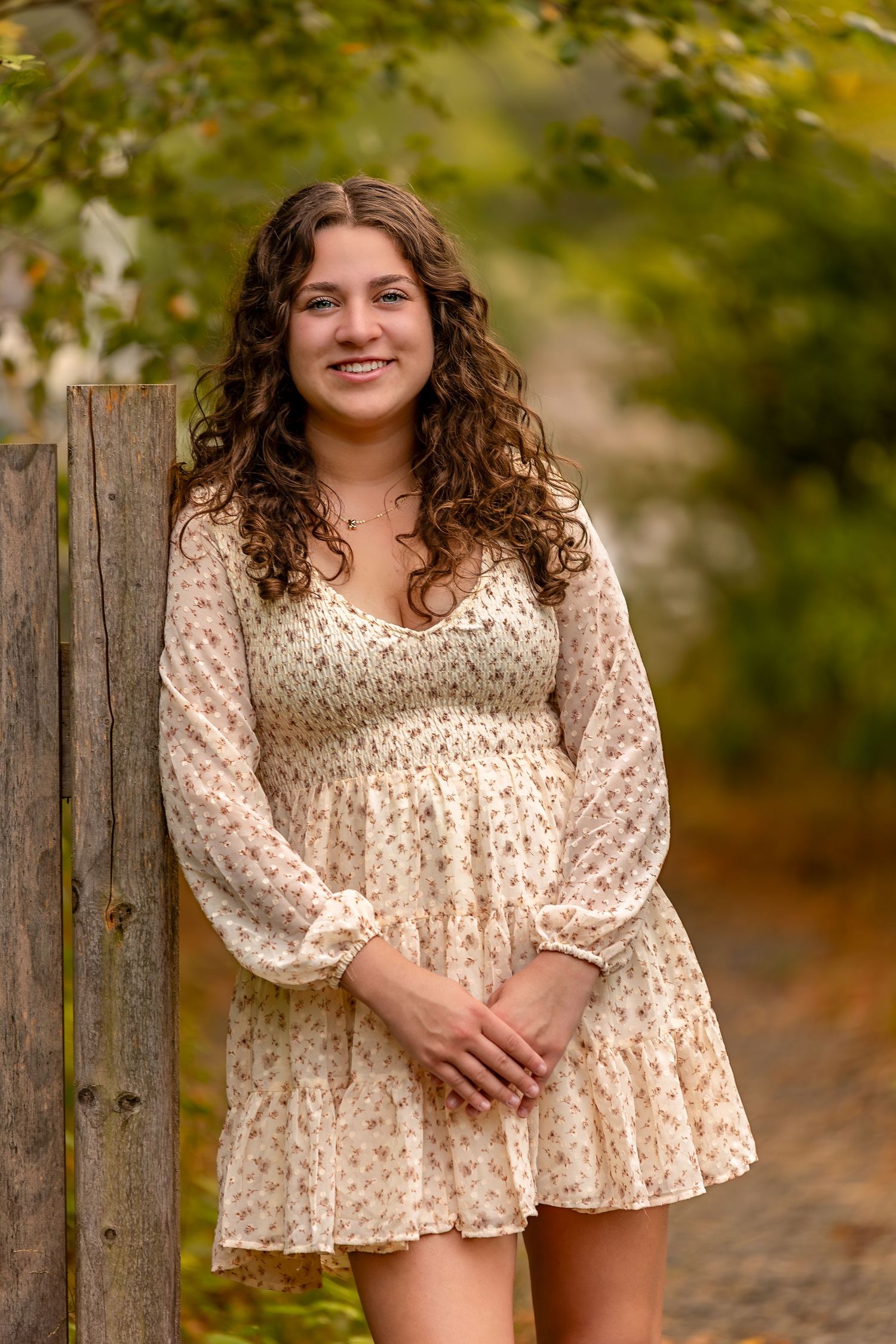 A graduating senior in cream floral dress poses outdoors against rustic wooden fence during autumn photoshoot.