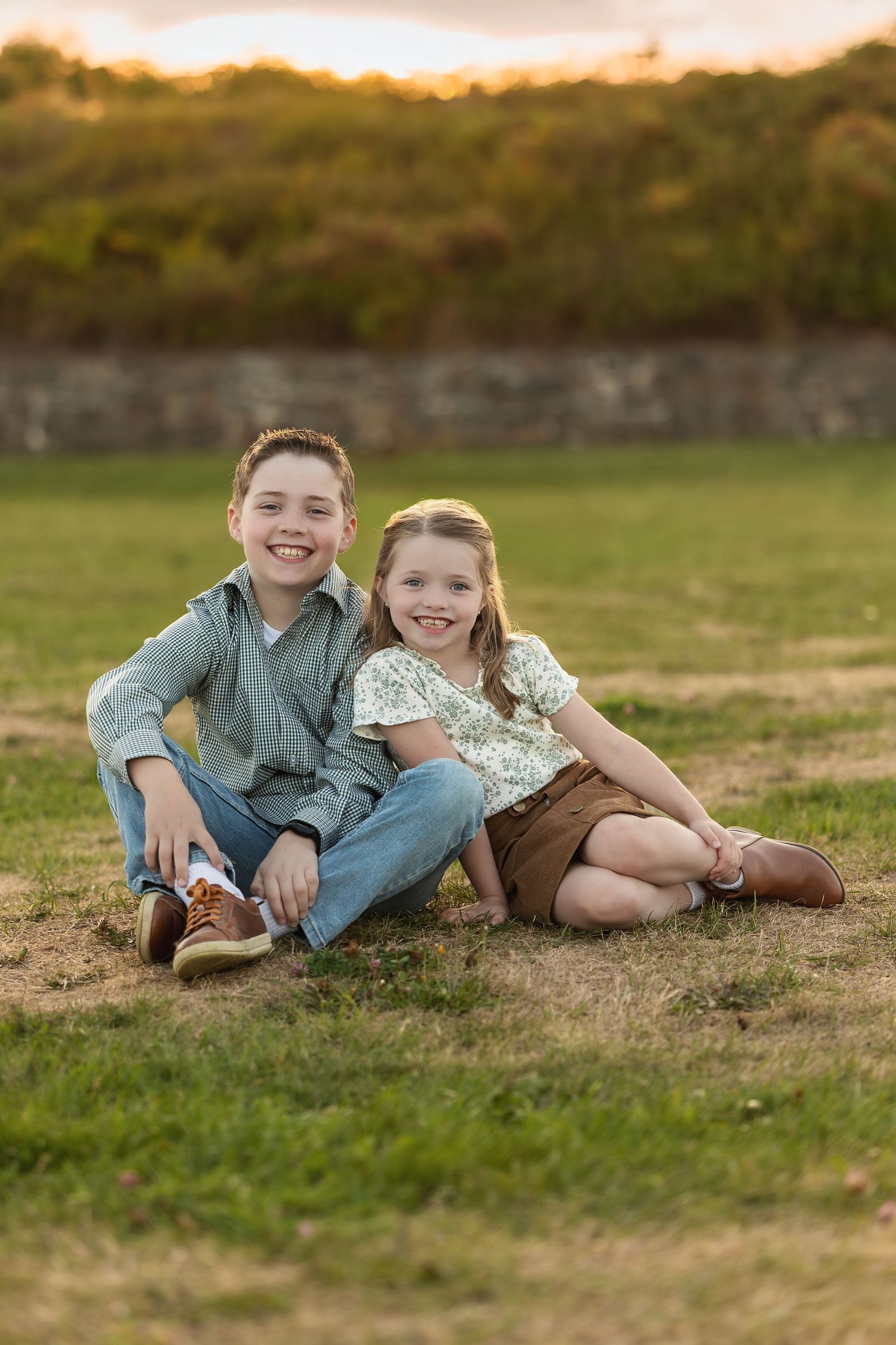 Two siblings sit together on grass during golden hour with natural backdrop.