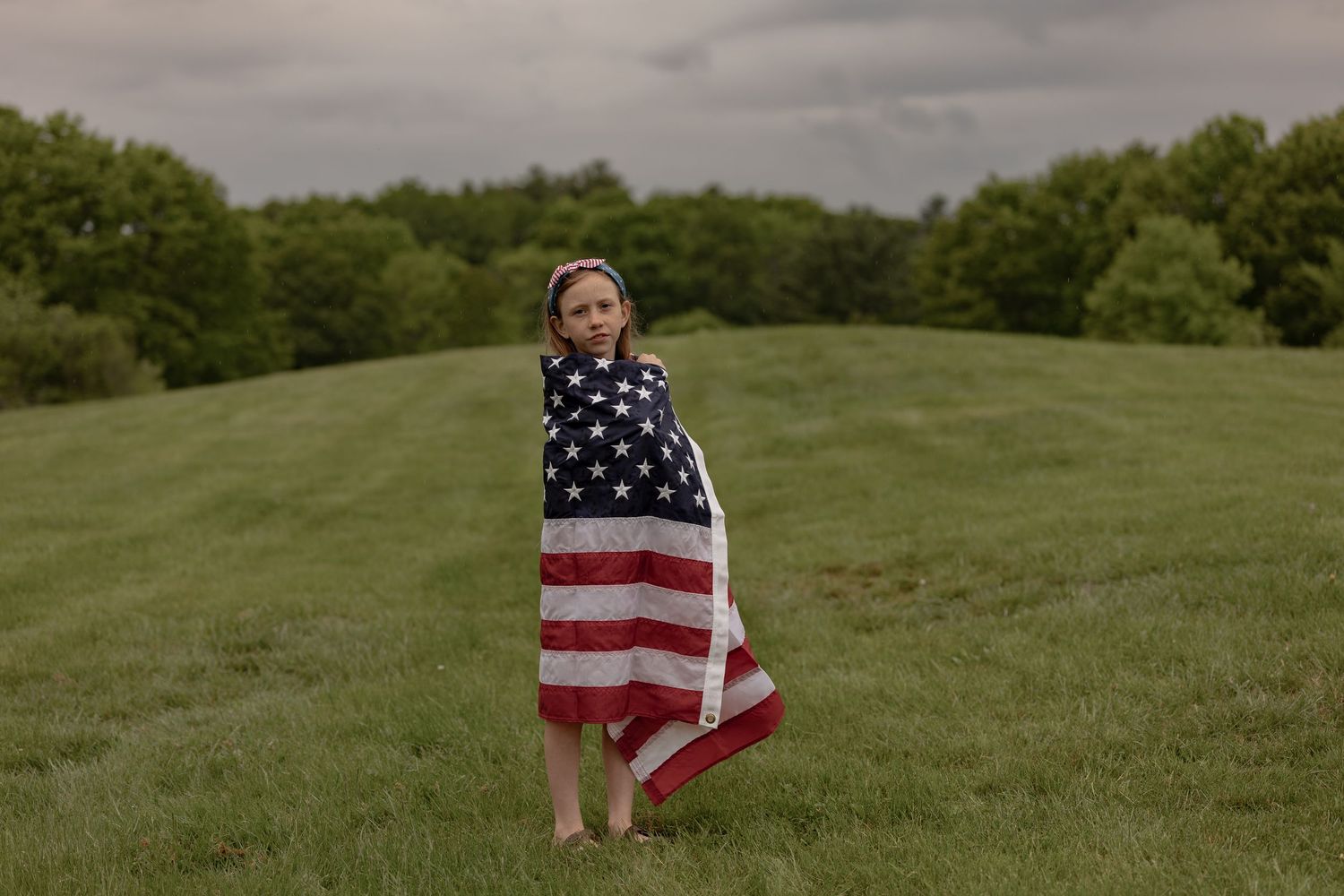 A barefoot child wrapped in an American flag stands alone on a lush green hillside under a cloudy sky.