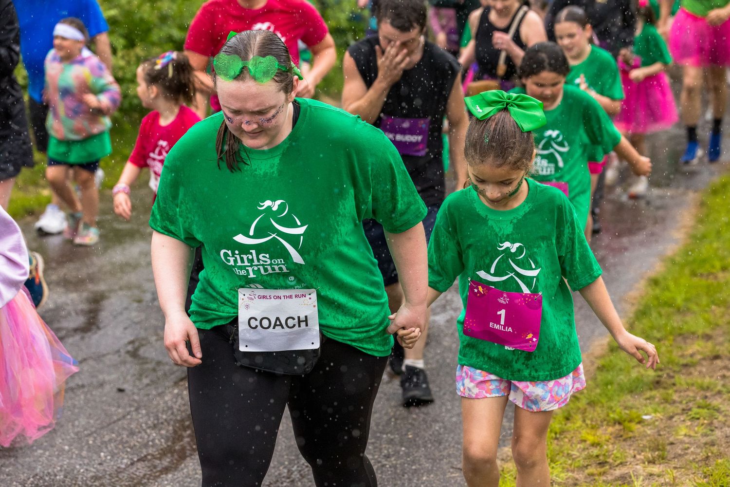 Girls on the Run coach in green shirt holds hands with young girl runner during a rainy charity 5K race event.