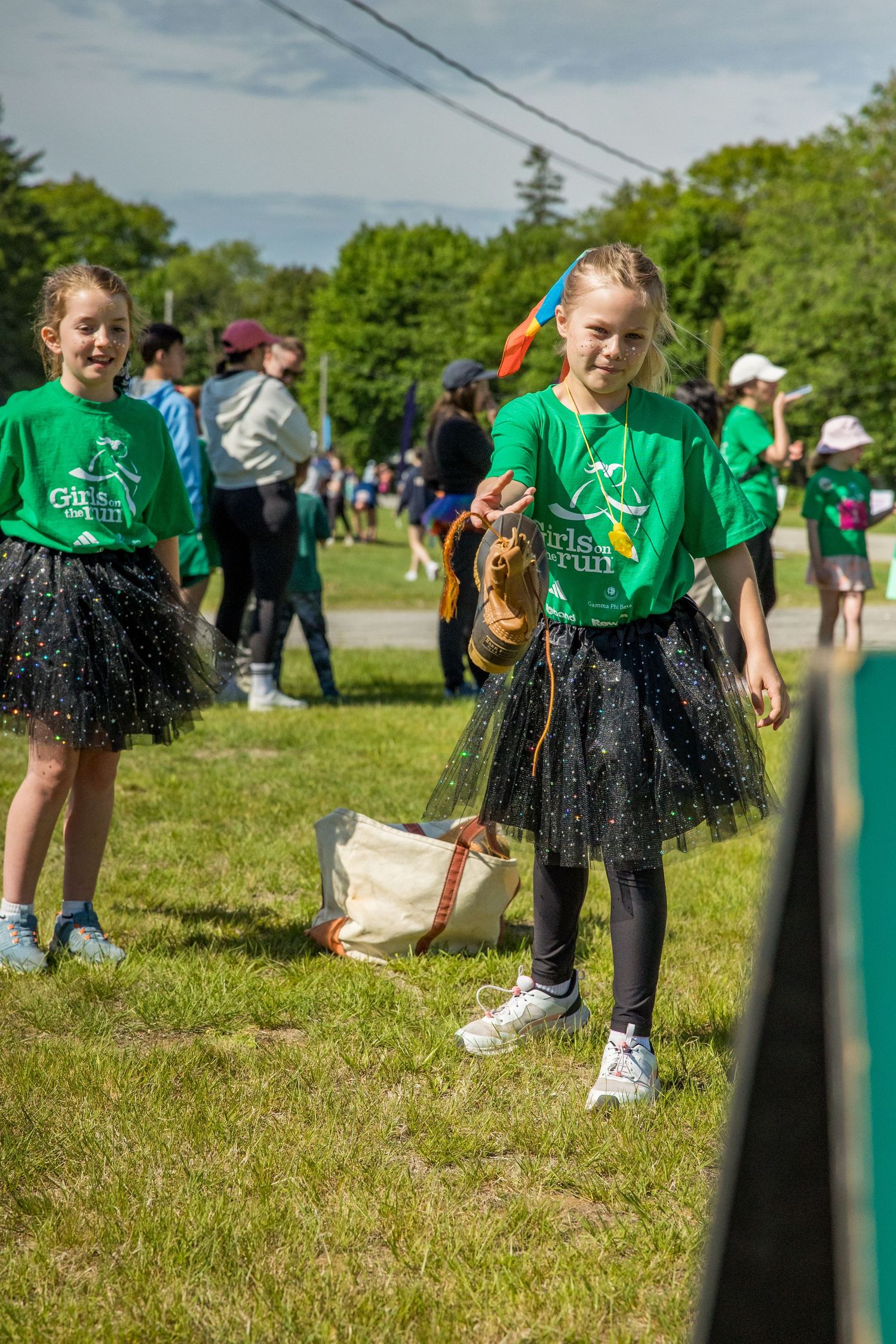 Two girls in green Girls on the Run shirts and black tutus stand outdoors at a running event on a sunny day.