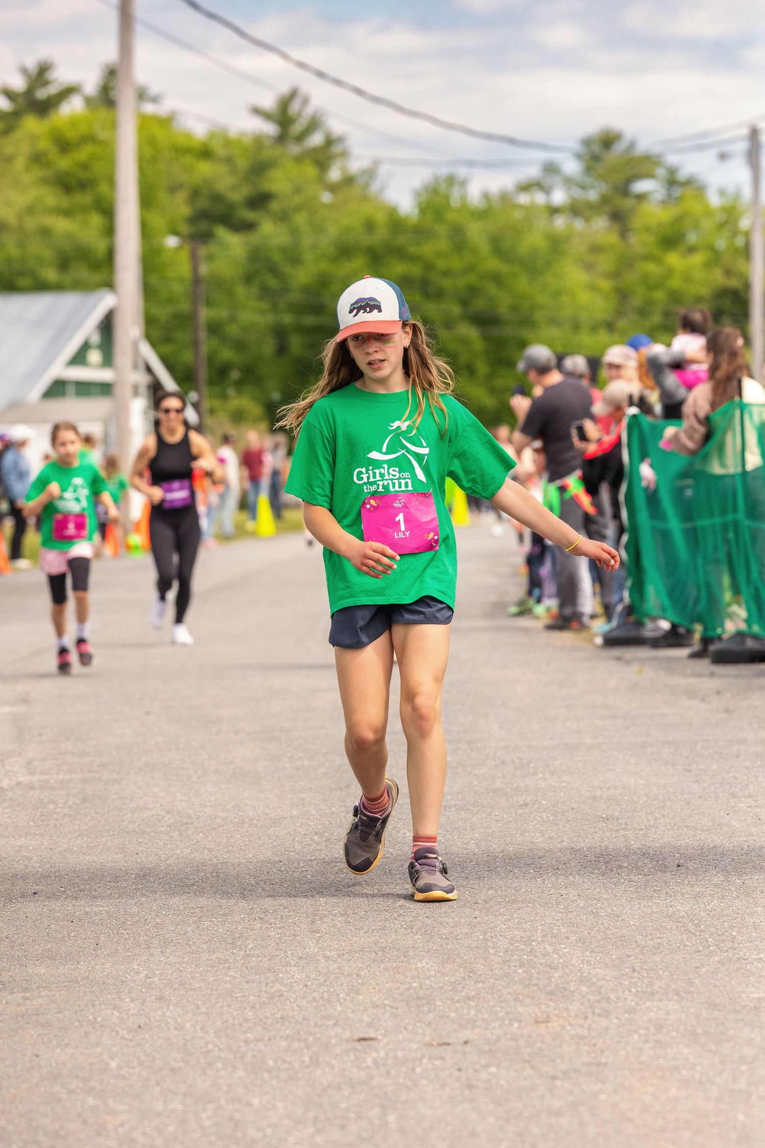 Young girl in green Girls on the Run shirt wearing bib number 1, running toward finish line at outdoor race event.