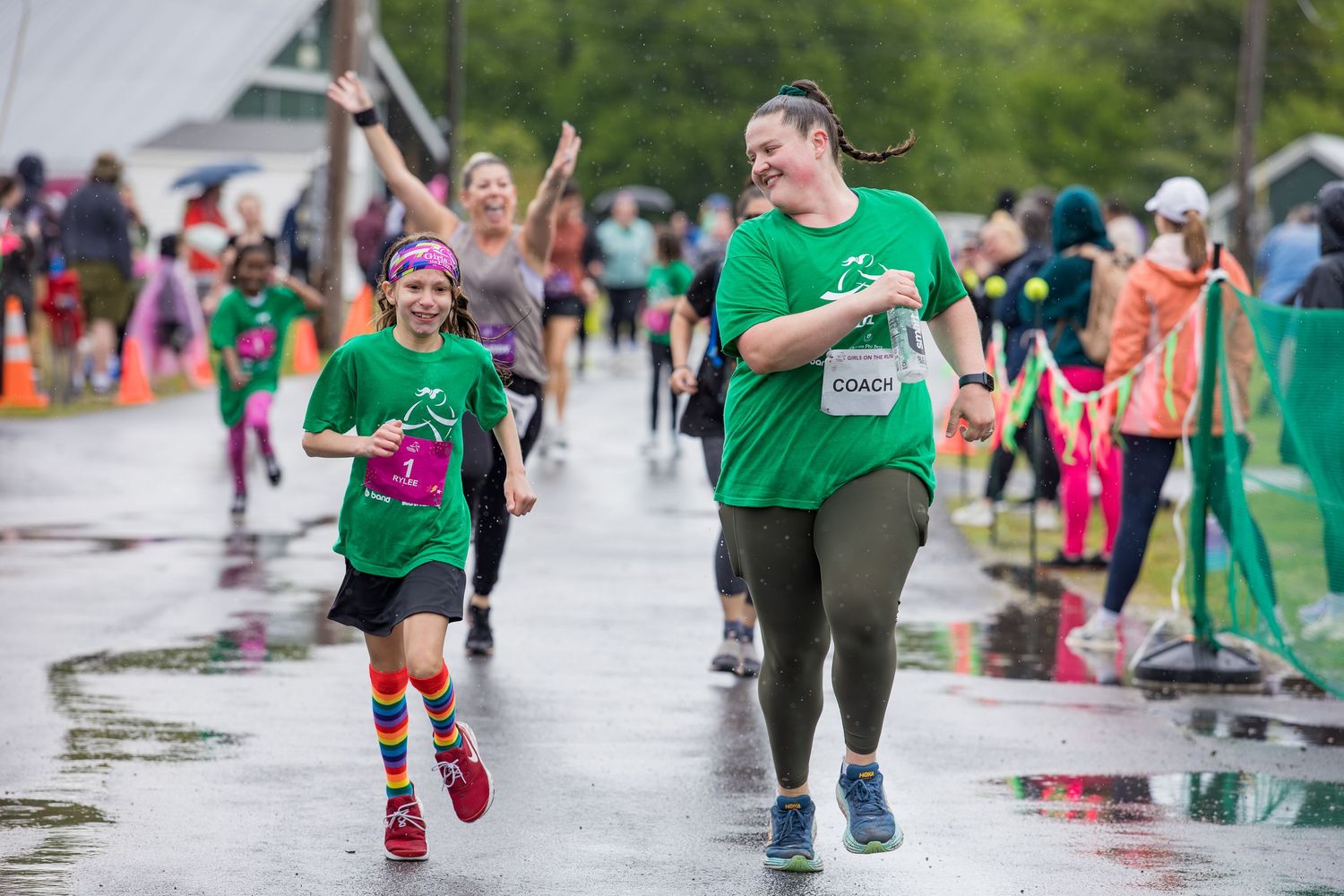 Woman coach and young girl in green shirts running together joyfully in a rainy outdoor fun run event.