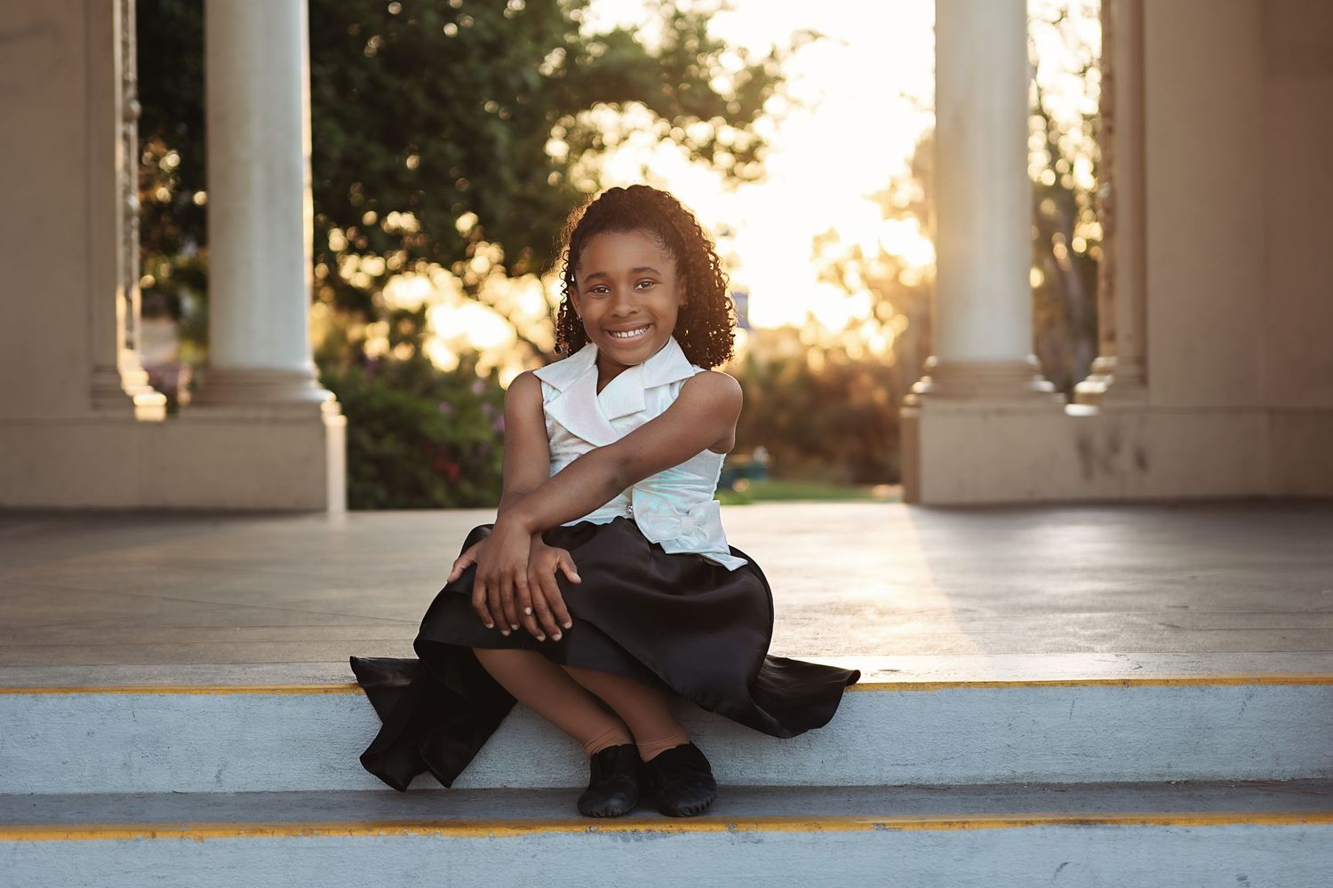Young girl in white top and black skirt sits smiling on steps with golden sunlight and columns behind her.