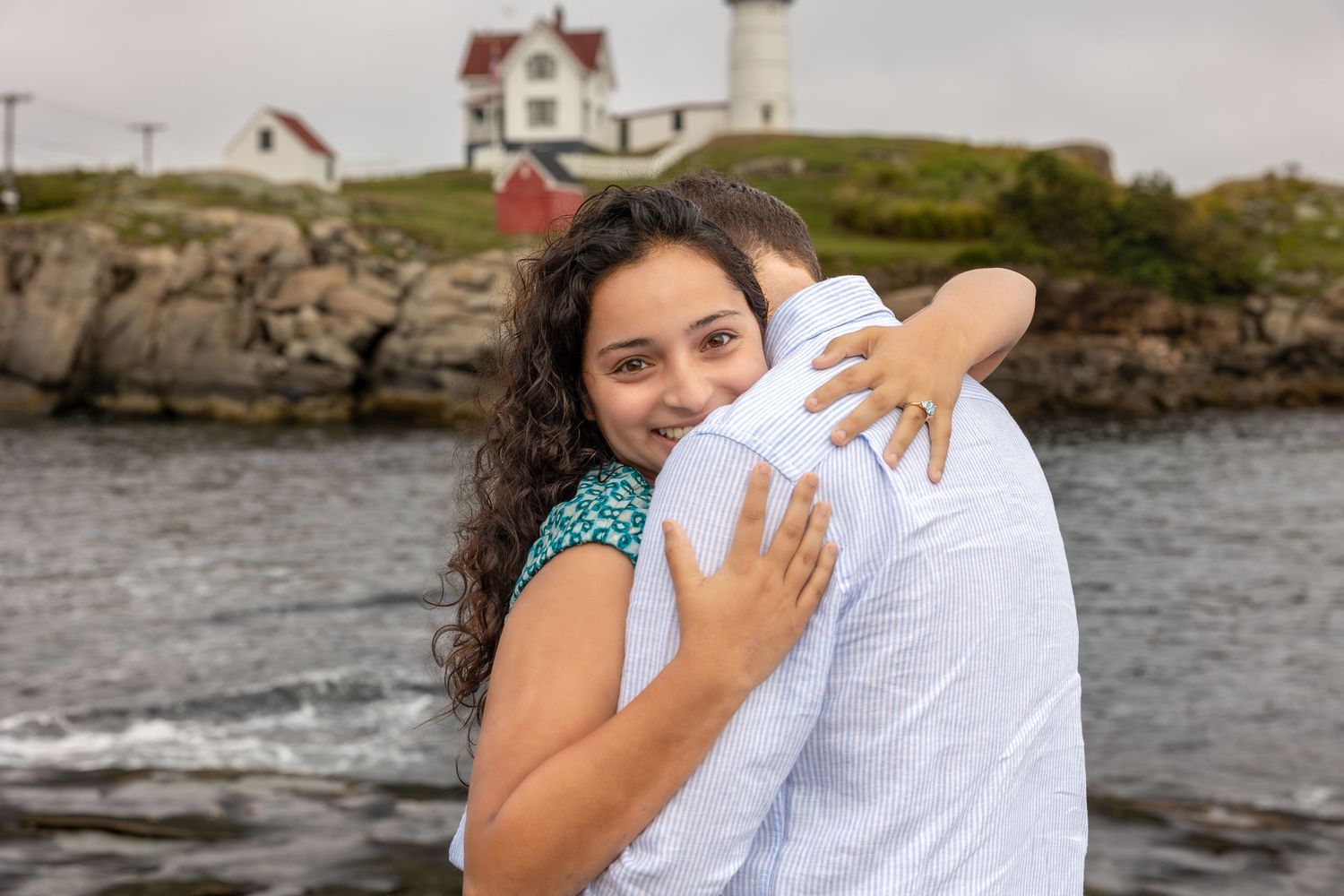 Couple embracing near rocky New England coastline with historic Nuble lighthouse in background.