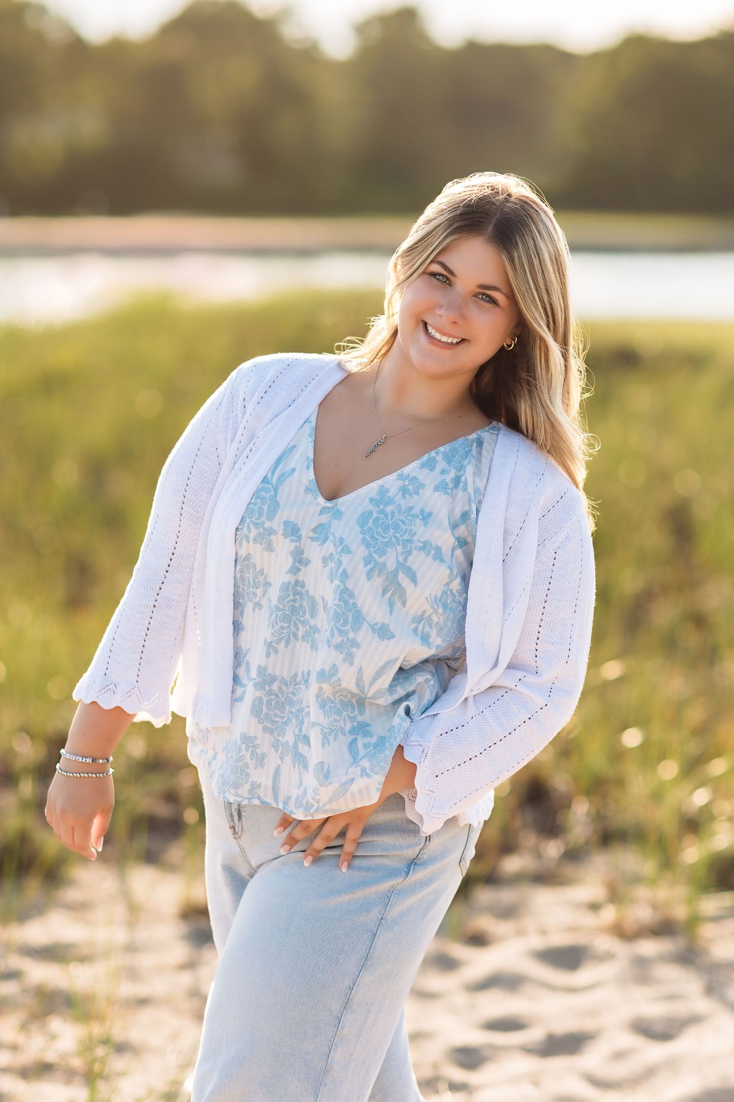 Smiling young woman in white cardigan and floral top poses on sandy beach path in golden hour light.